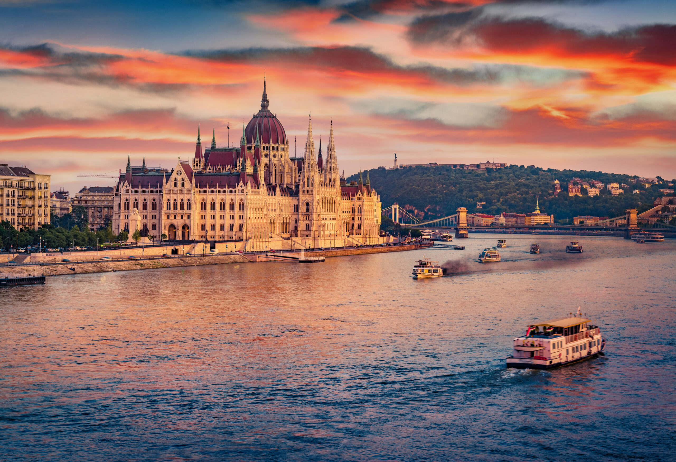 Budapest Parliament Building along the Danube at sunset with boats on the water and a pink-orange sky.