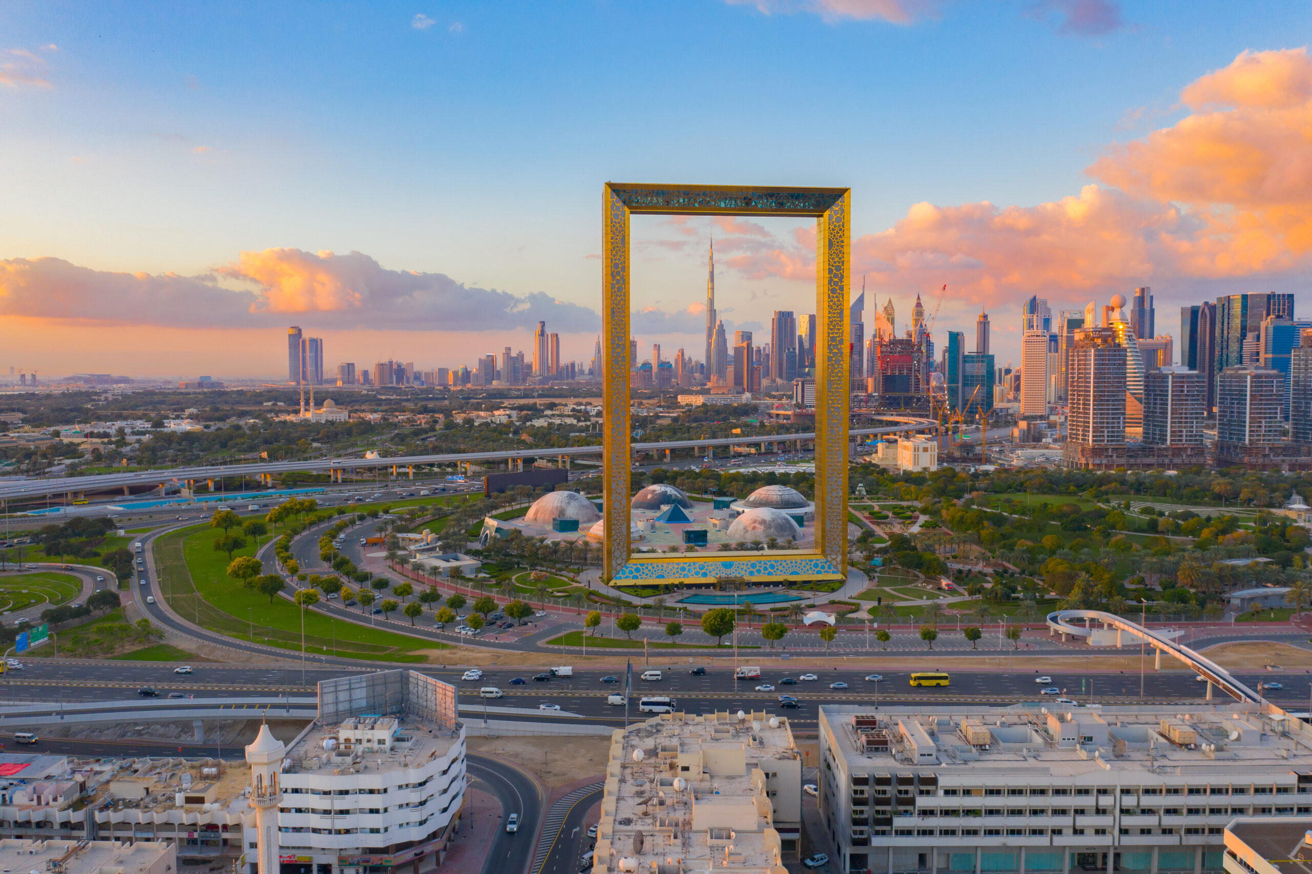 Dubai Frame frames the city skyline at sunset, with skyscrapers in the background and a warm, orange-pink sky.