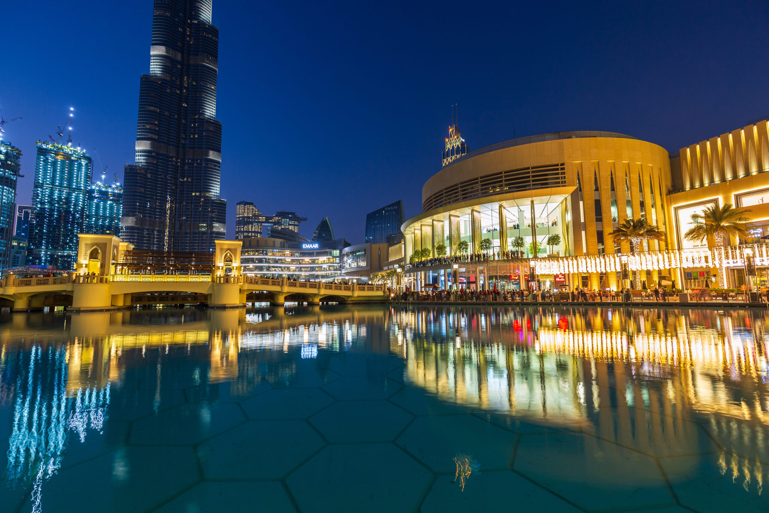 Night view of Dubai skyline with Burj Khalifa, illuminated shopping complex, and reflections on calm water.