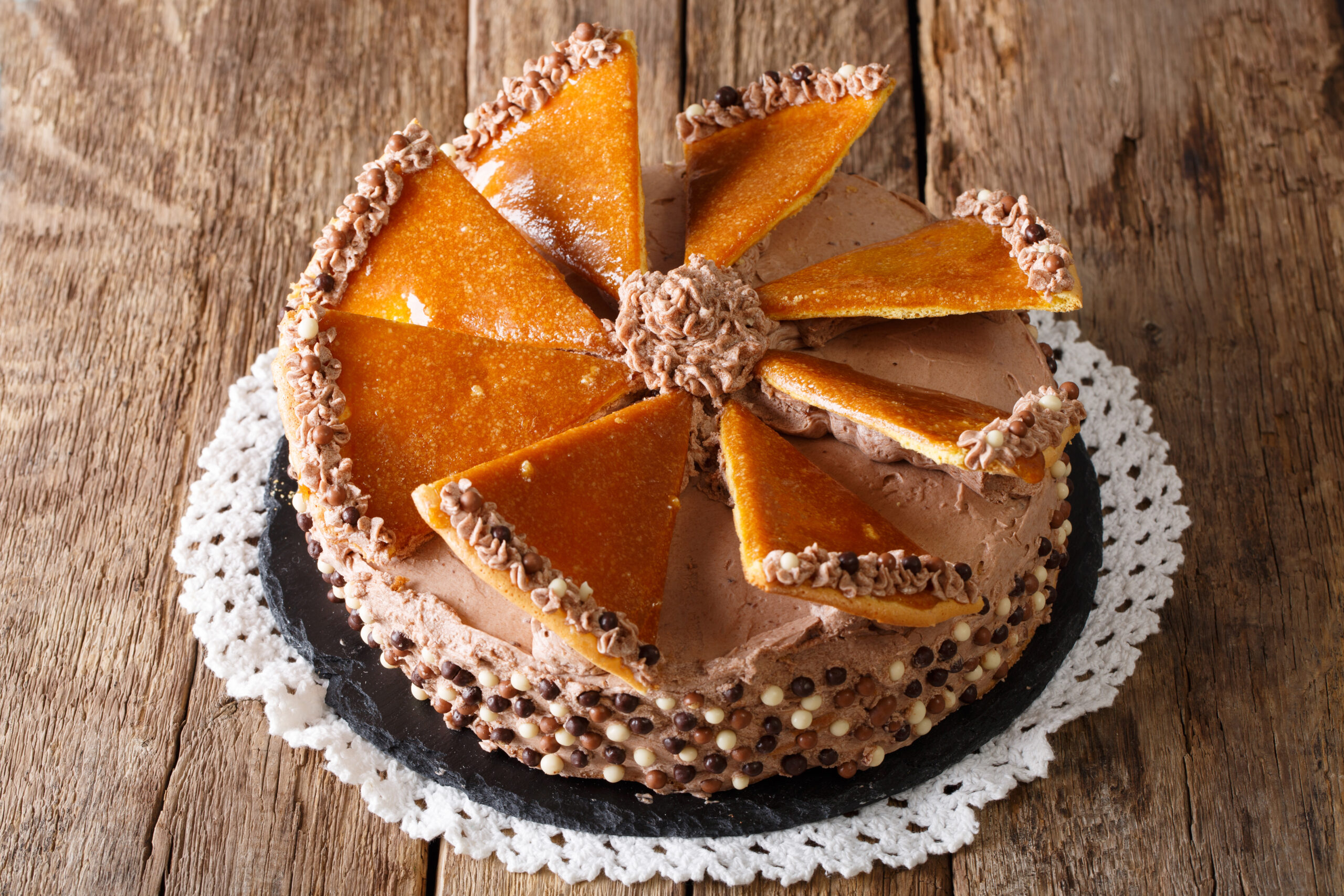 Cake with orange segments arranged around the edge, chocolate frosting, and chocolate pearls on a wooden table with a lace doily underneath.