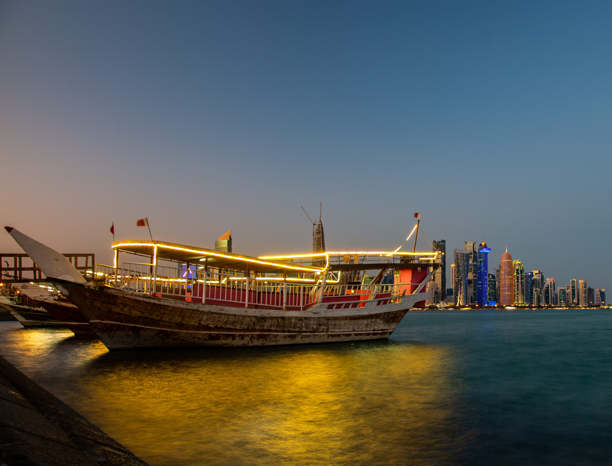 Nighttime harbor with illuminated traditional boats on calm water and a skyline of tall buildings in the distance.