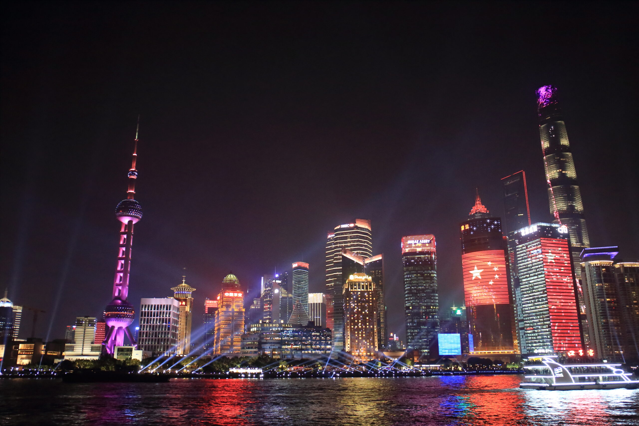 Night skyline of a illuminated city along a river, with pink/purple skyscraper lights reflecting on the water