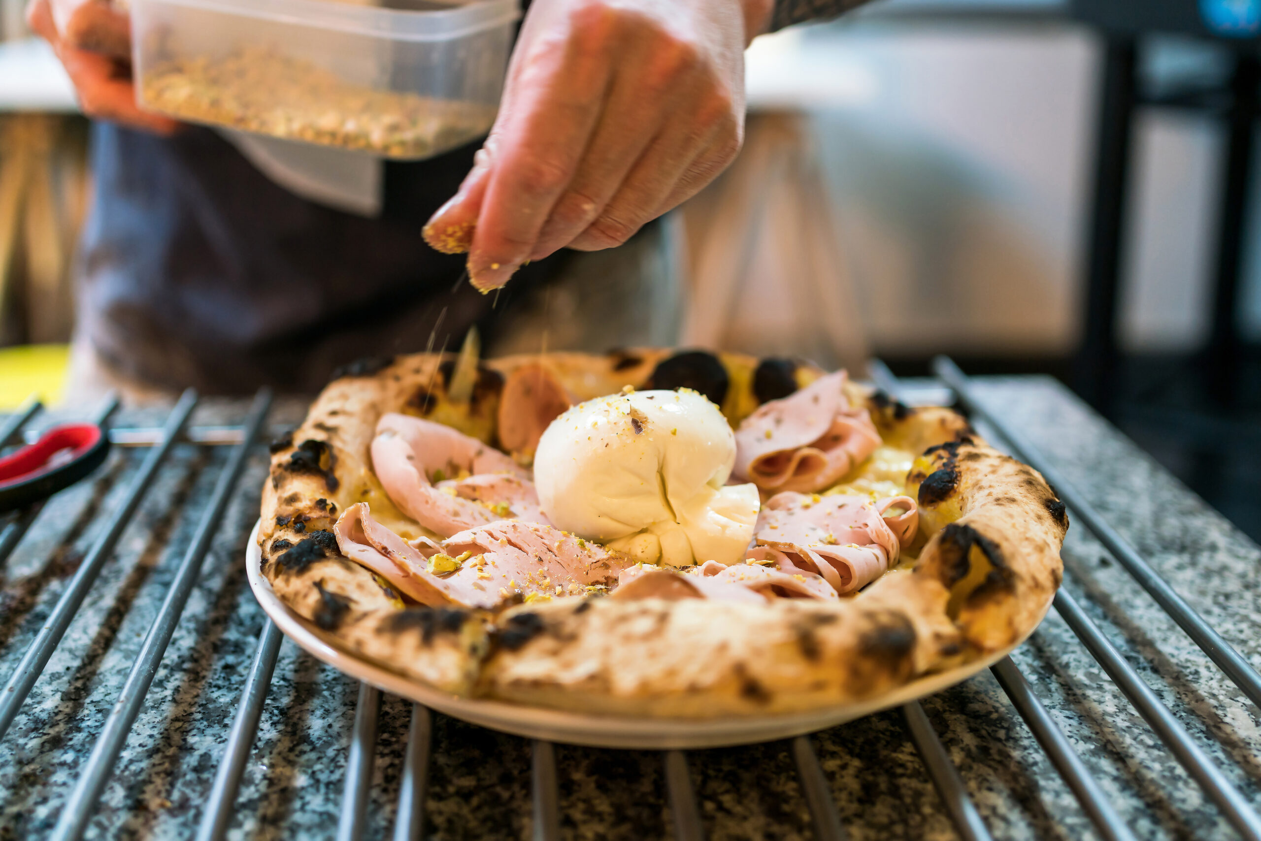 Close-up of a cook sprinkling herbs and spices over a ham-and-m mozzarella pizza on a grill rack.