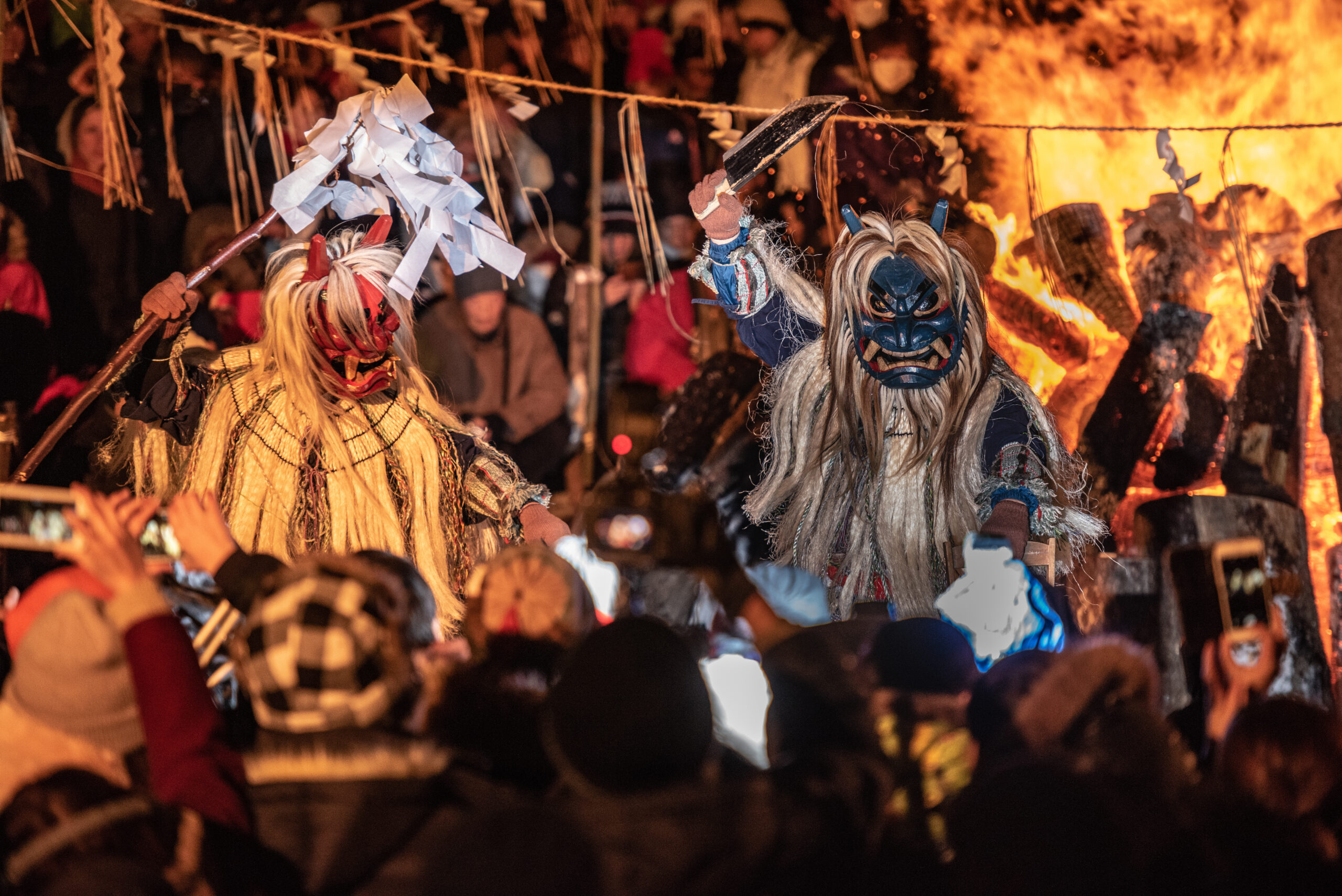Masked performers in colorful costumes perform before a roaring bonfire as a crowd watches at night festival.