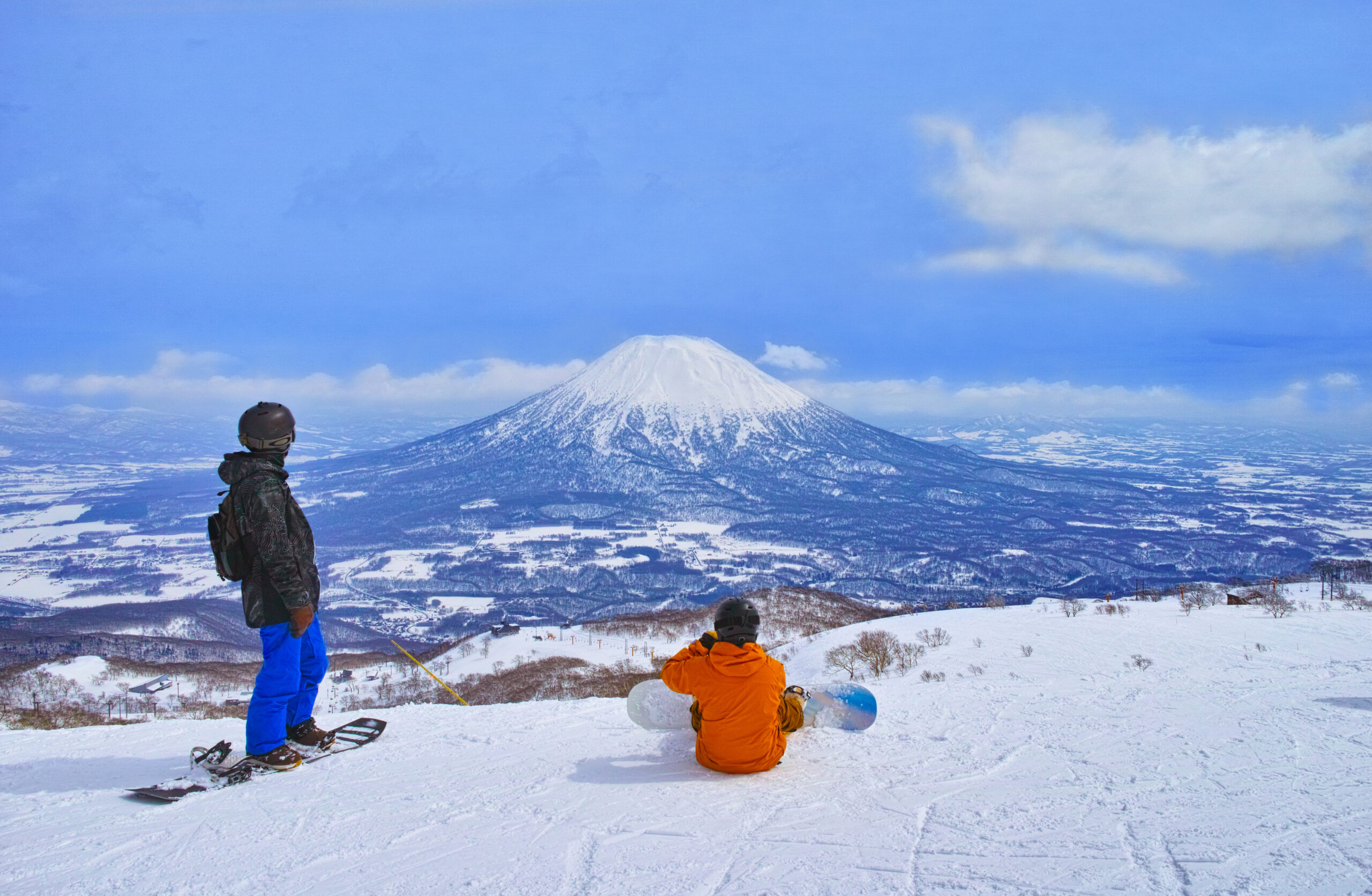 Two snowboarders overlook a snow-covered valley with a large snowy mountain in the distance; one stands left in blue pants and a helmet, another sits in an orange jacket with a snowboard nearby.