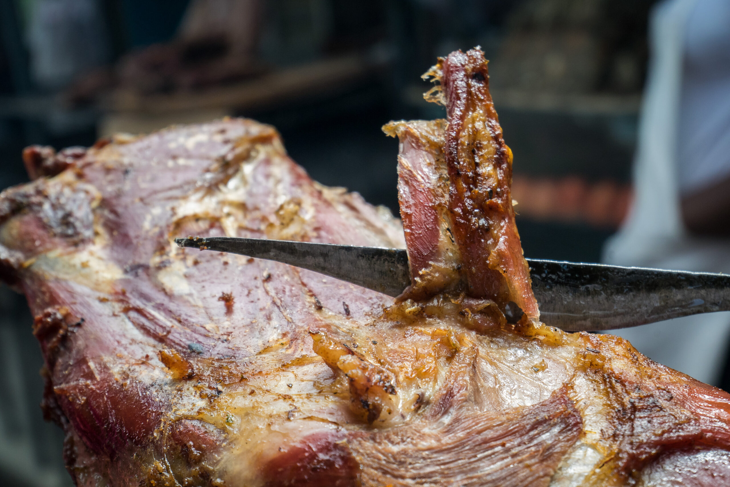Close-up of a large piece of smoked meat on a grill, with a knife slicing through a strip of meat and a browned, glazed crust visible.