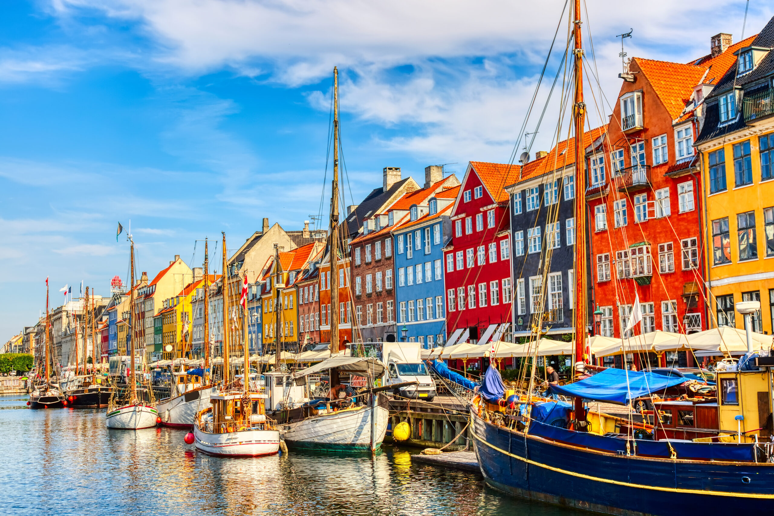 Colorful row houses along a harbor with moored boats and calm water under a blue sky, likely Nyhavn Copenhagen street scene.