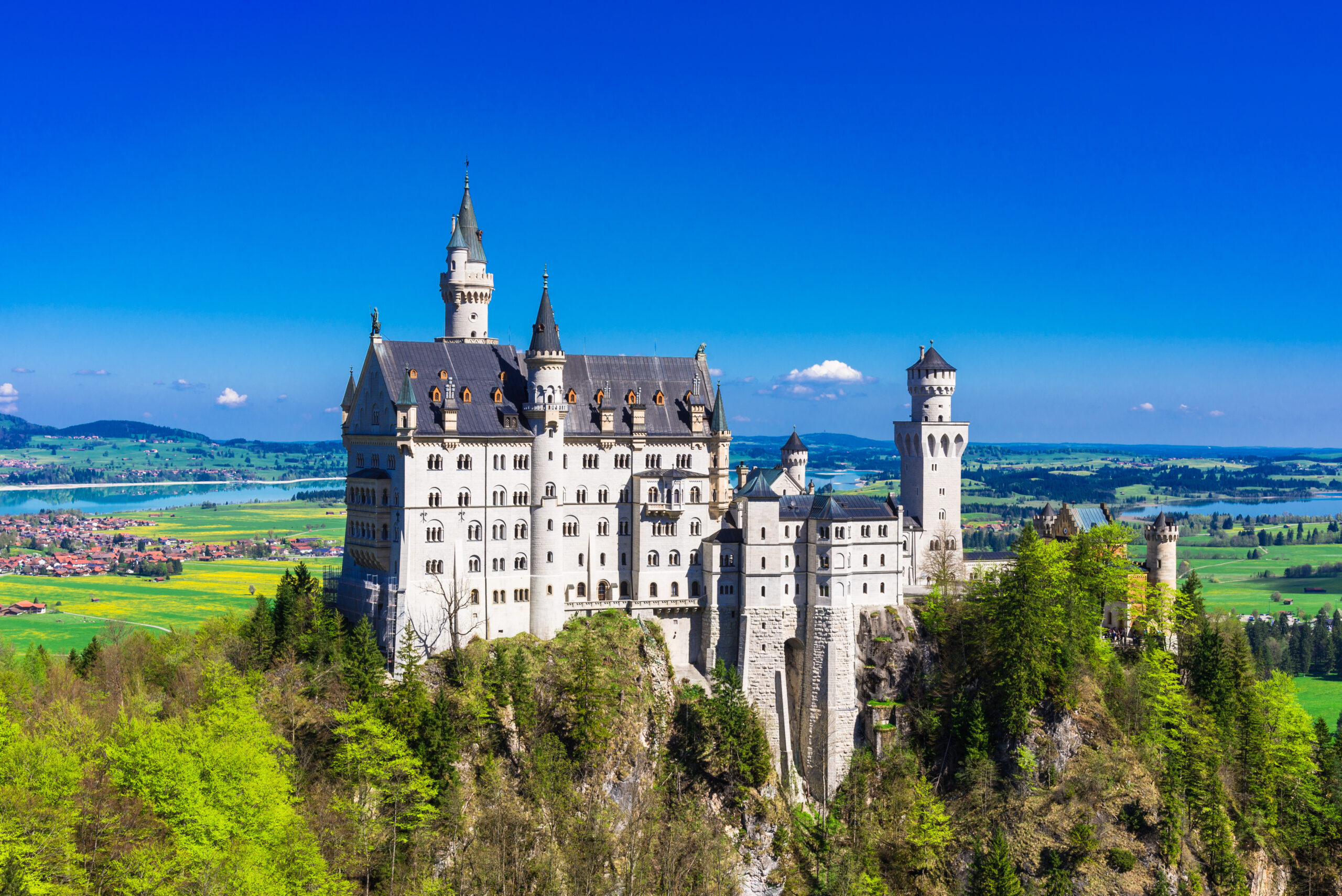 Castle with tall spires atop a rocky cliff, surrounded by green forest and a valley beneath the bright blue sky.