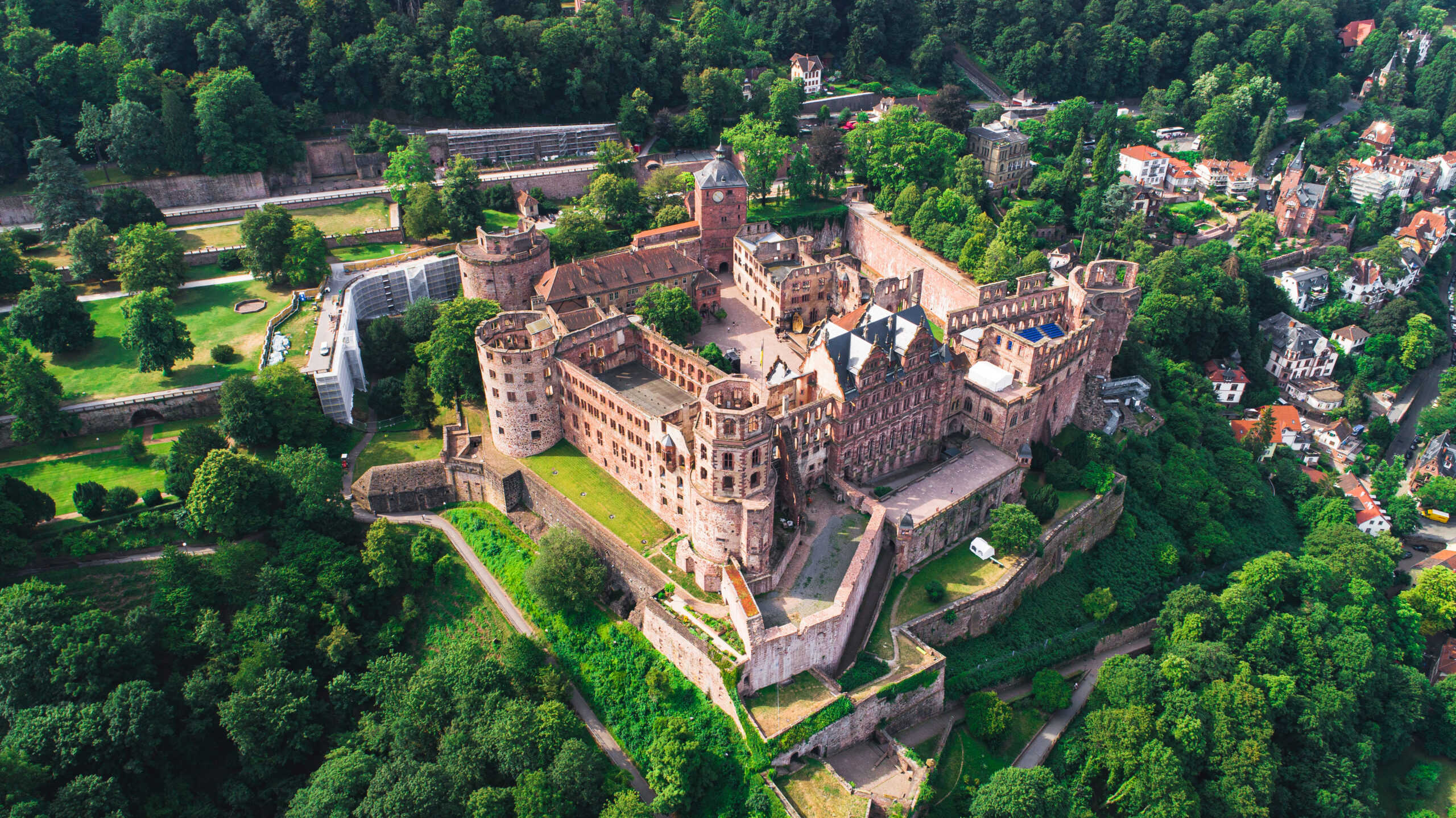 Aerial view of a historic brick castle with towers and walls, set among lush green trees and a nearby village.