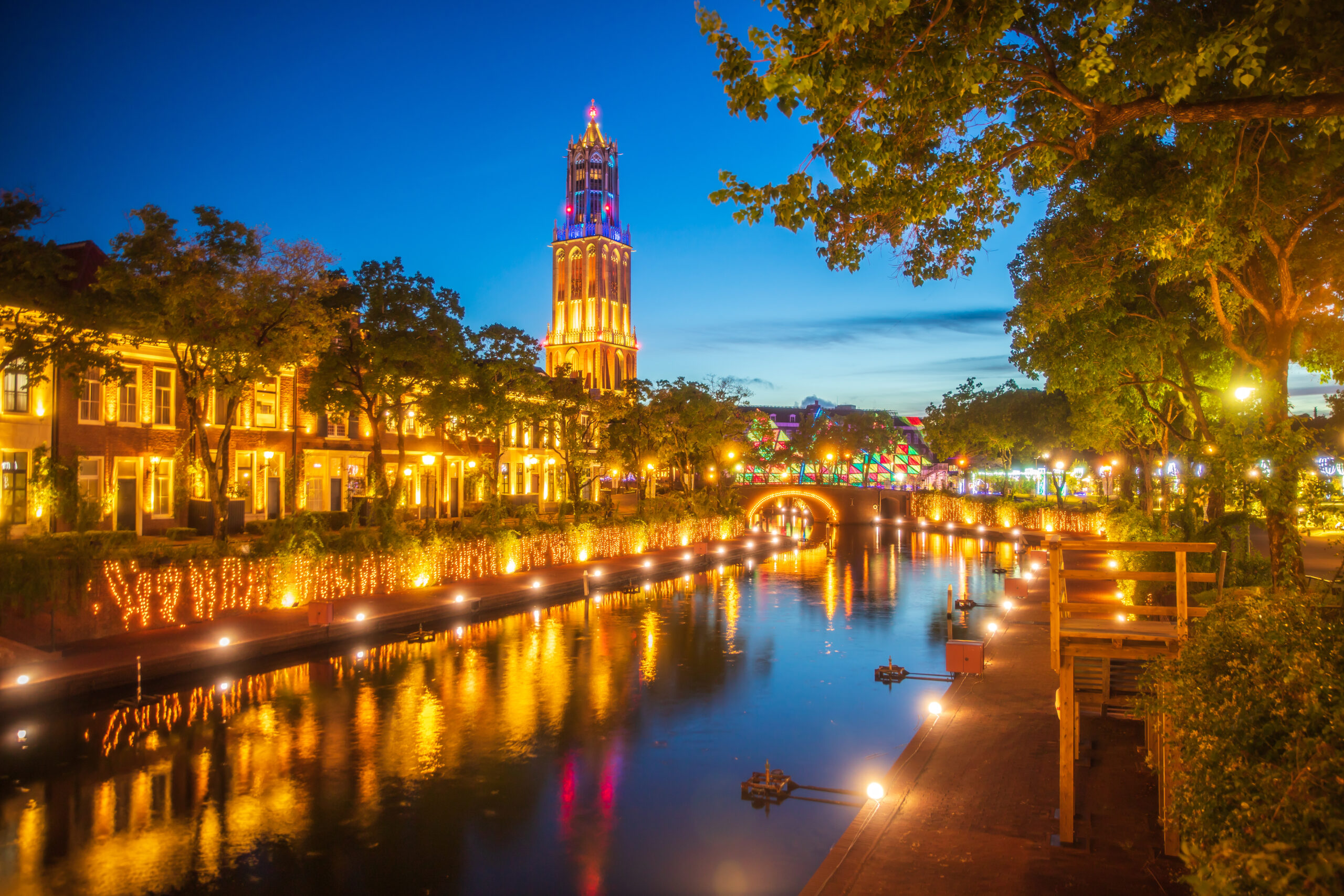 Night cityscape: canal with warm-lit buildings, trees, and a tall, glowing tower at center reflecting in the water.
