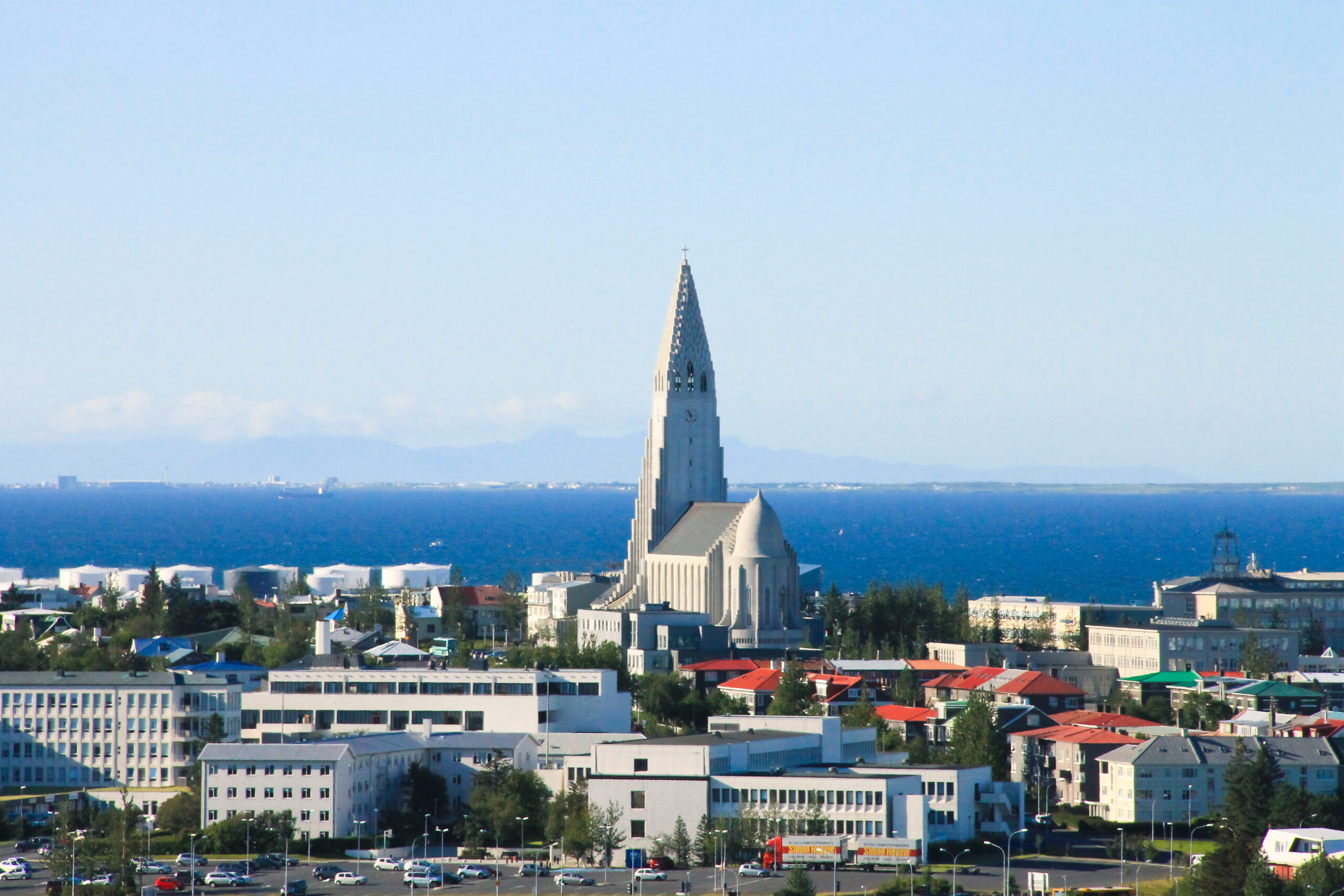 Prominent white tower with a pointed spire rising above a coastal city skyline by the sea.