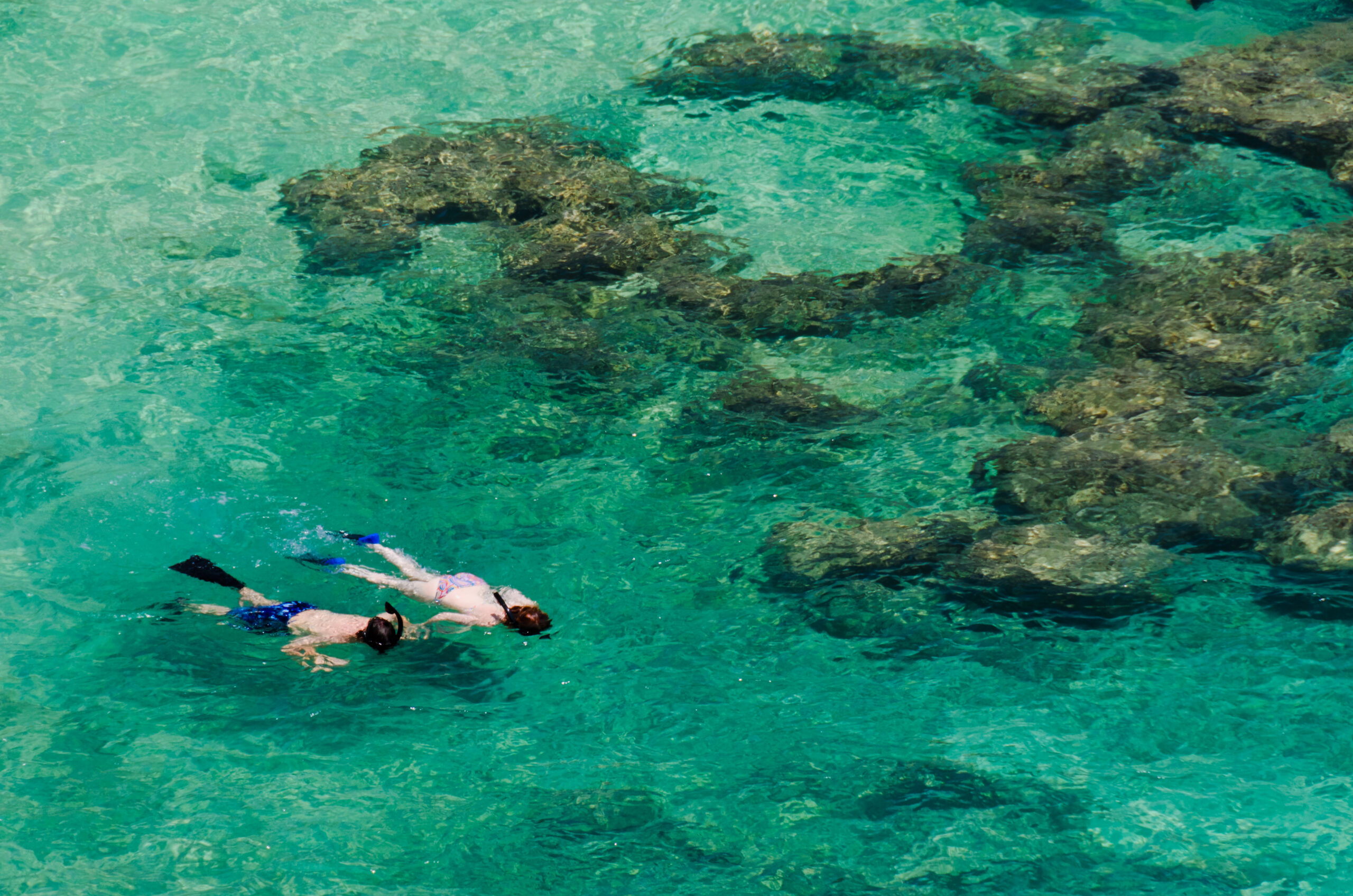 Two snorkelers with fins swim above a rocky reef in clear turquoise water.