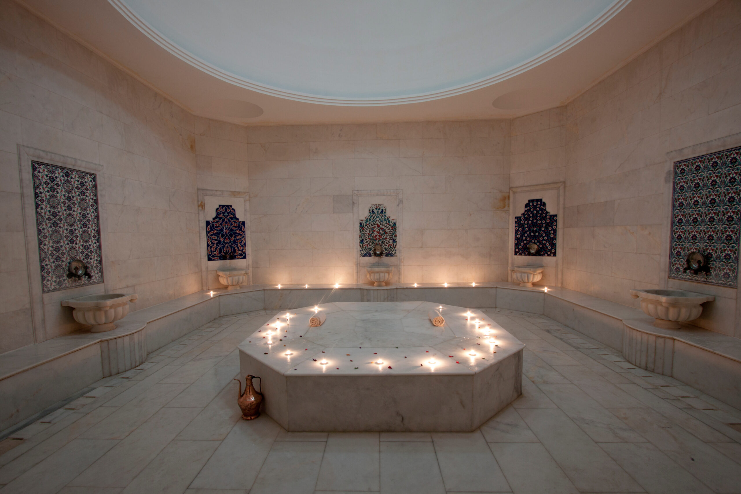 Marble interior of a spa bath with alcoves featuring decorative ceramic tiles and candles around a central hexagonal pool.