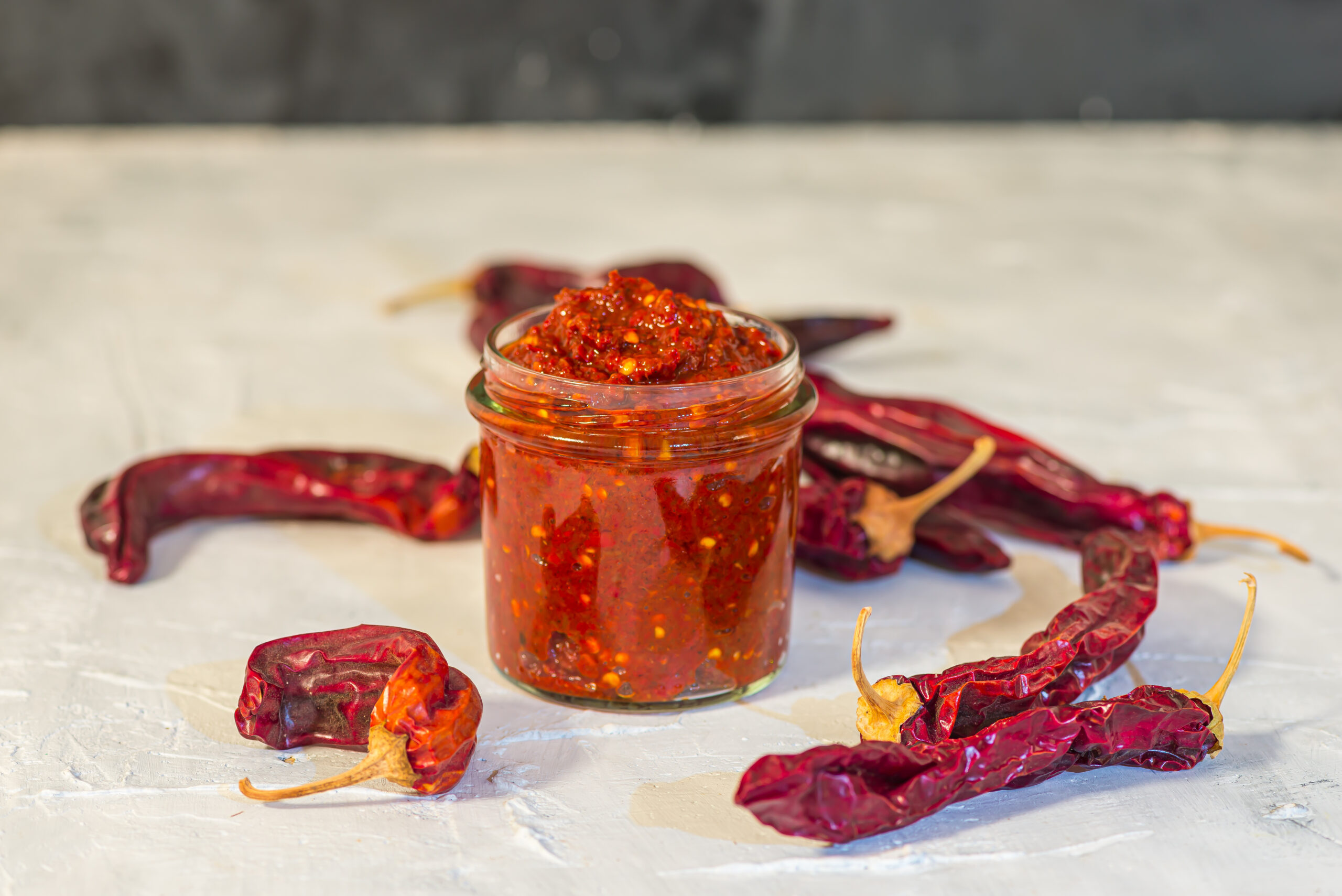 Glass jar of red chili paste with chili flakes, surrounded by dried red chilies on a light surface