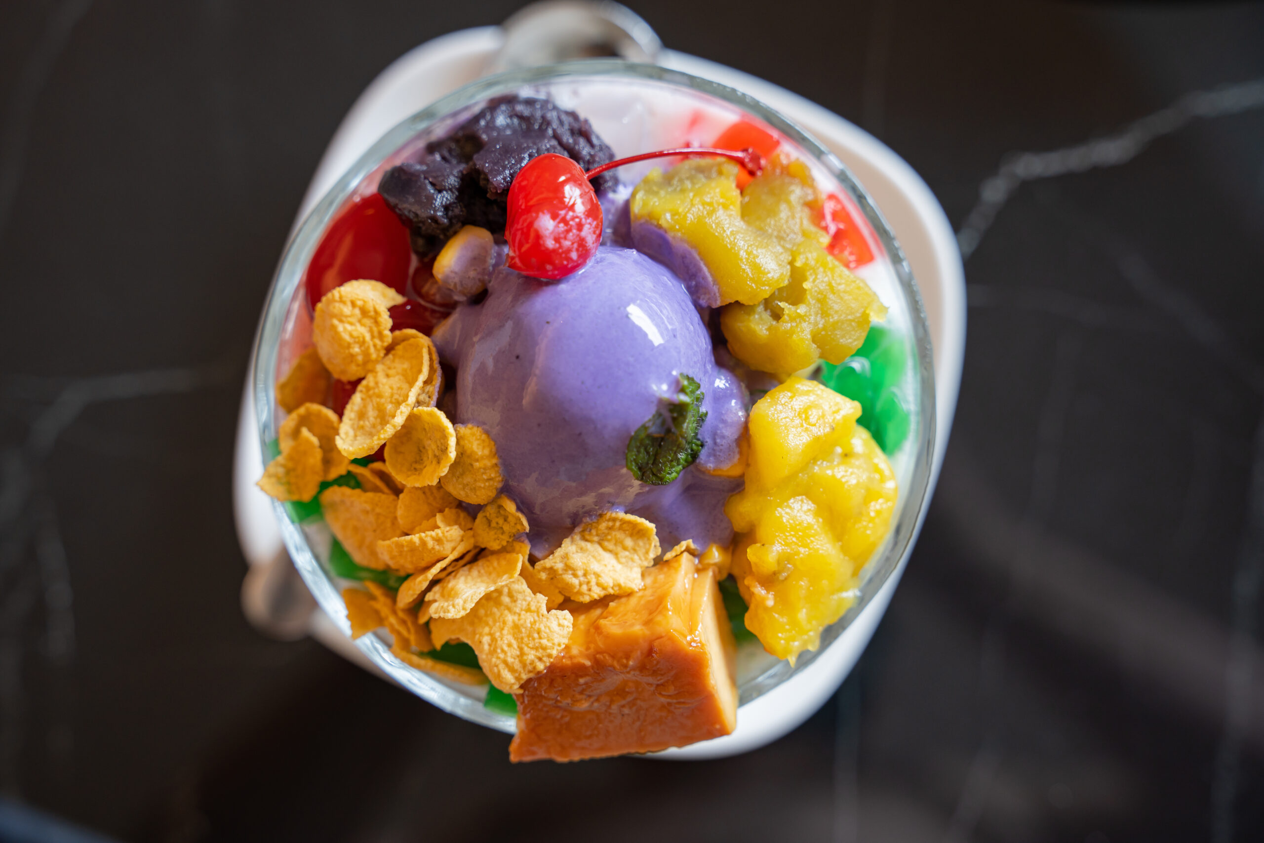 Colorful ice cream sundae in a glass: purple scoop with mango chunks, cornflakes, a cherry, and a pastry piece on top