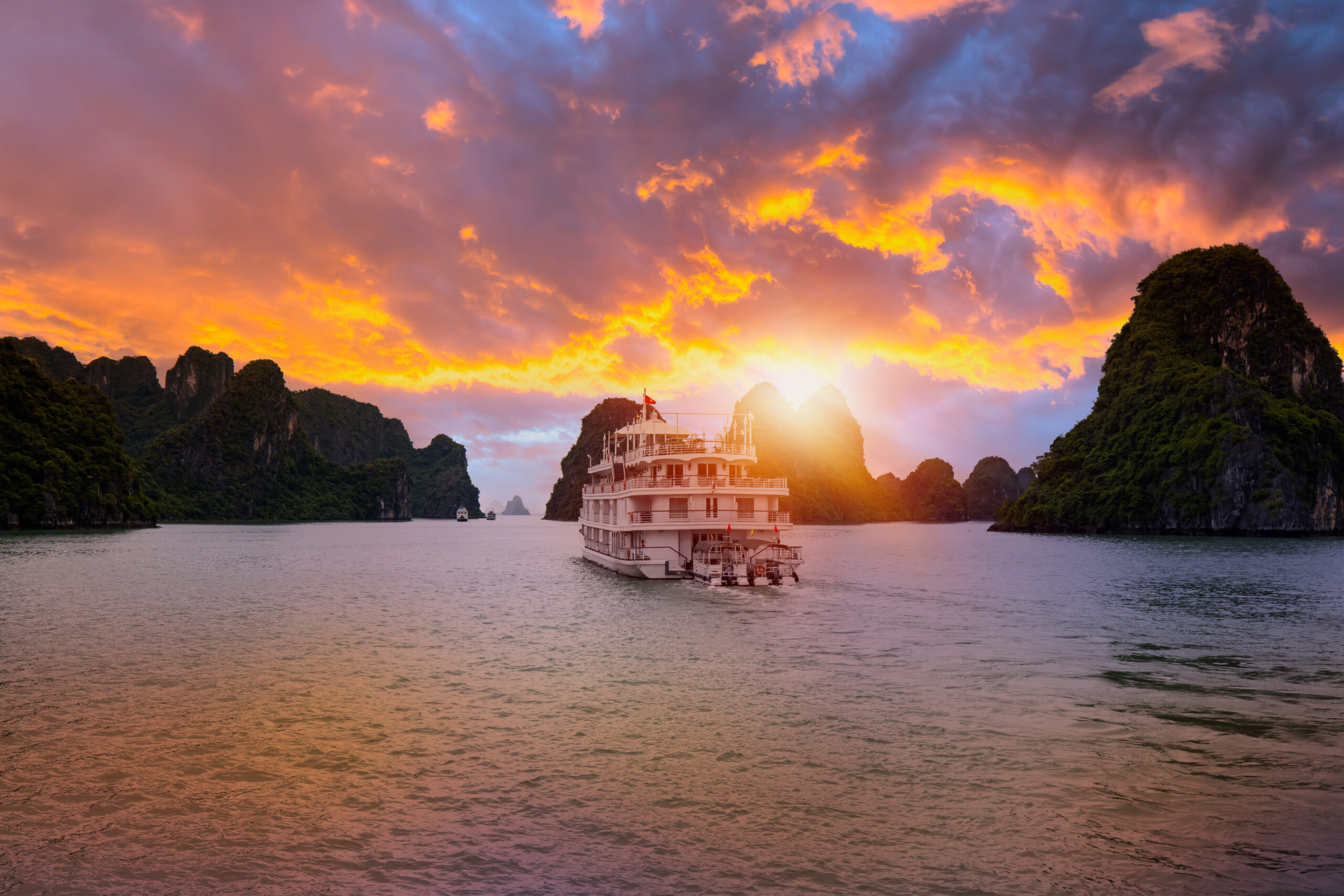 White passenger cruise ship on calm bay at sunset, with an orange–purple sky and lush limestone islands in the background.