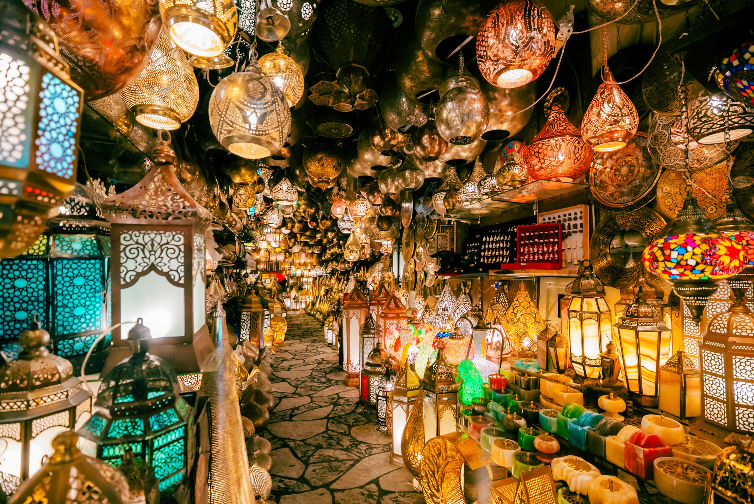 A vibrant market stall densely lit with ornate metal and glass lanterns hanging from the ceiling and lining the walkway, glowing warm colors.