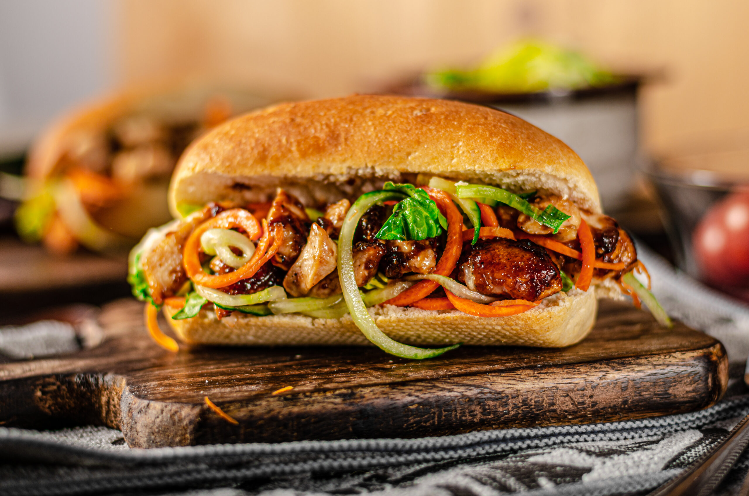 Close-up of a sandwich on a toasted bun filled with caramelized meat, shredded carrots, onions, and greens on a rustic wooden board.