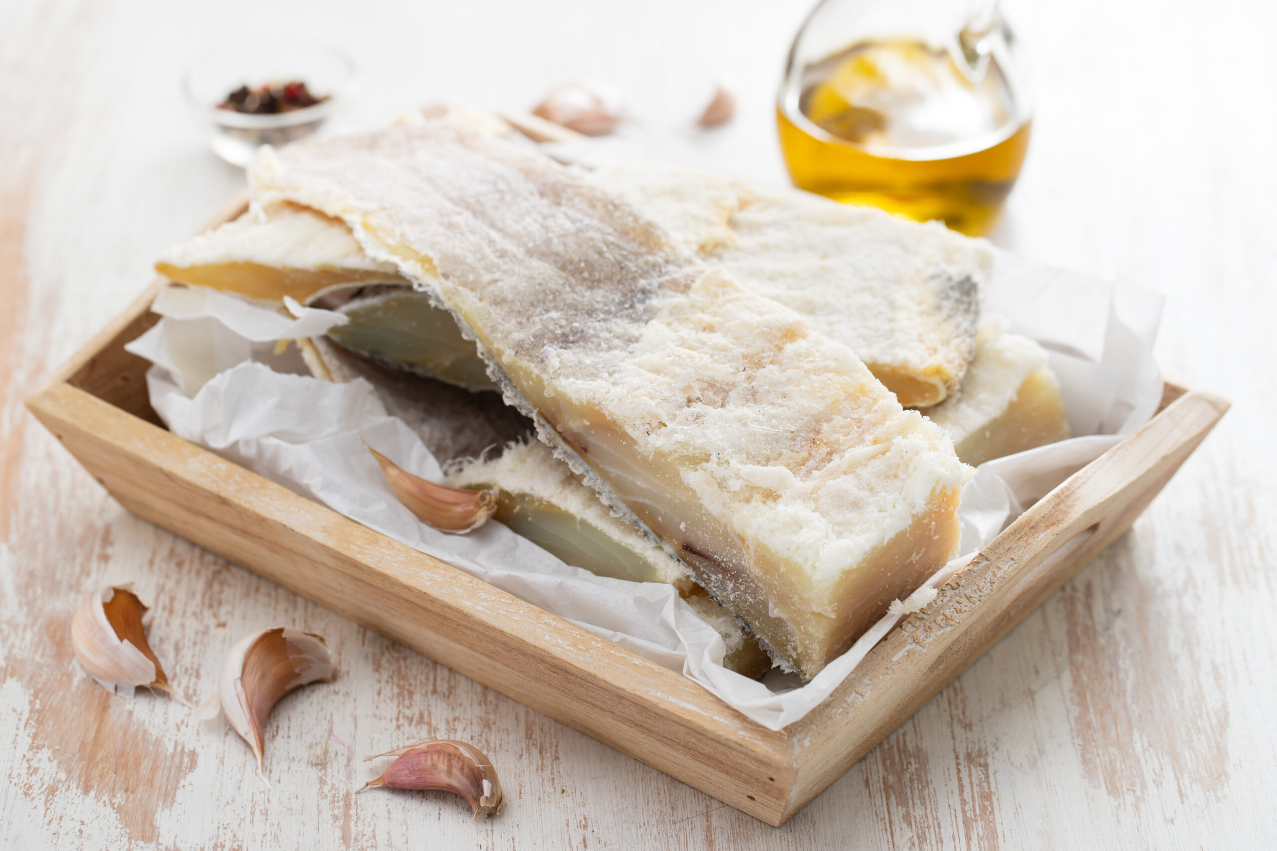 Salted cod fillets in a wooden crate, dusted with salt, with garlic cloves and a bottle of oil in the background.