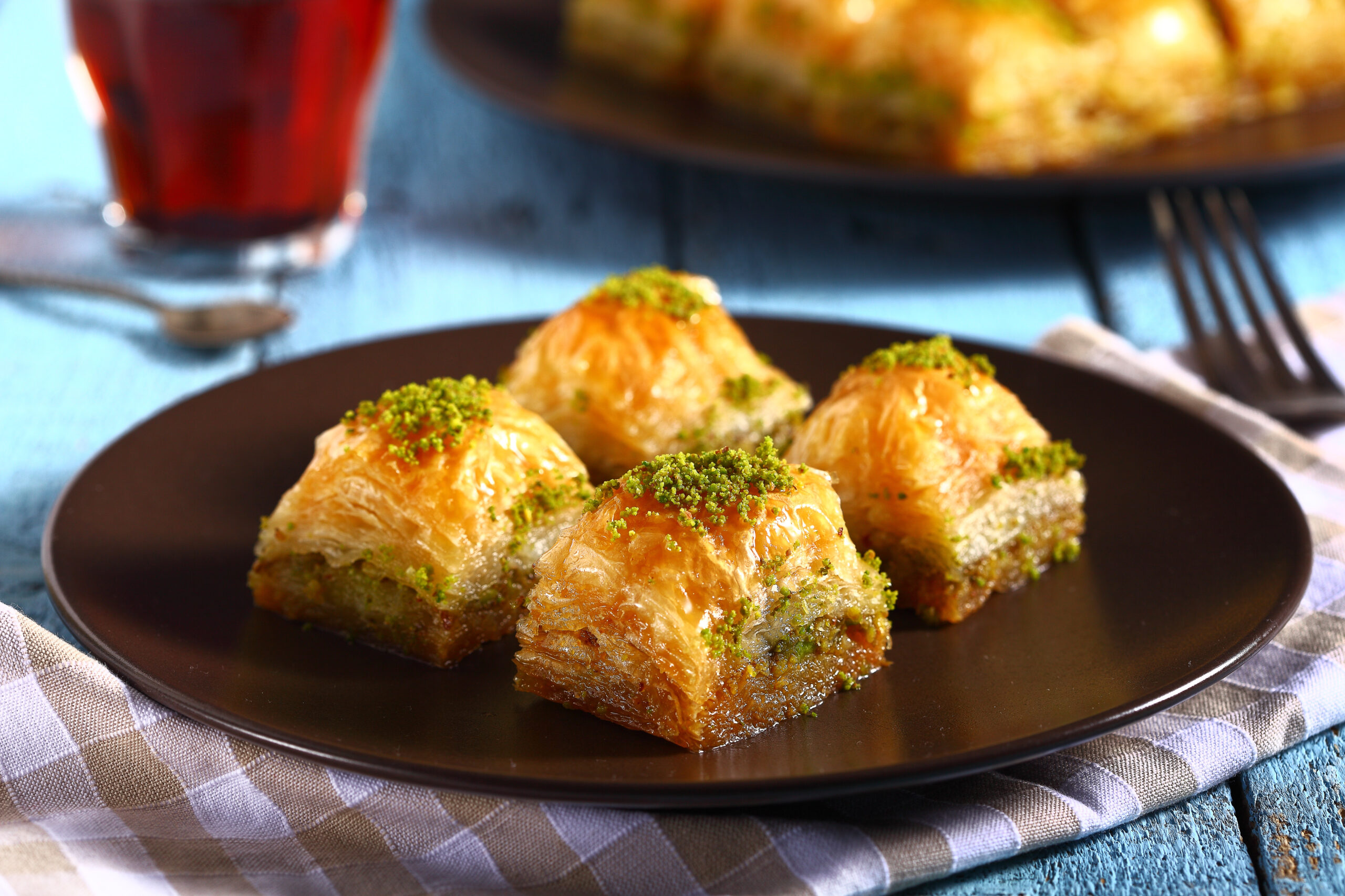 Flaky pastry bites topped with green pistachios on a dark plate, blue tablecloth beneath, glass of red drink in the background.