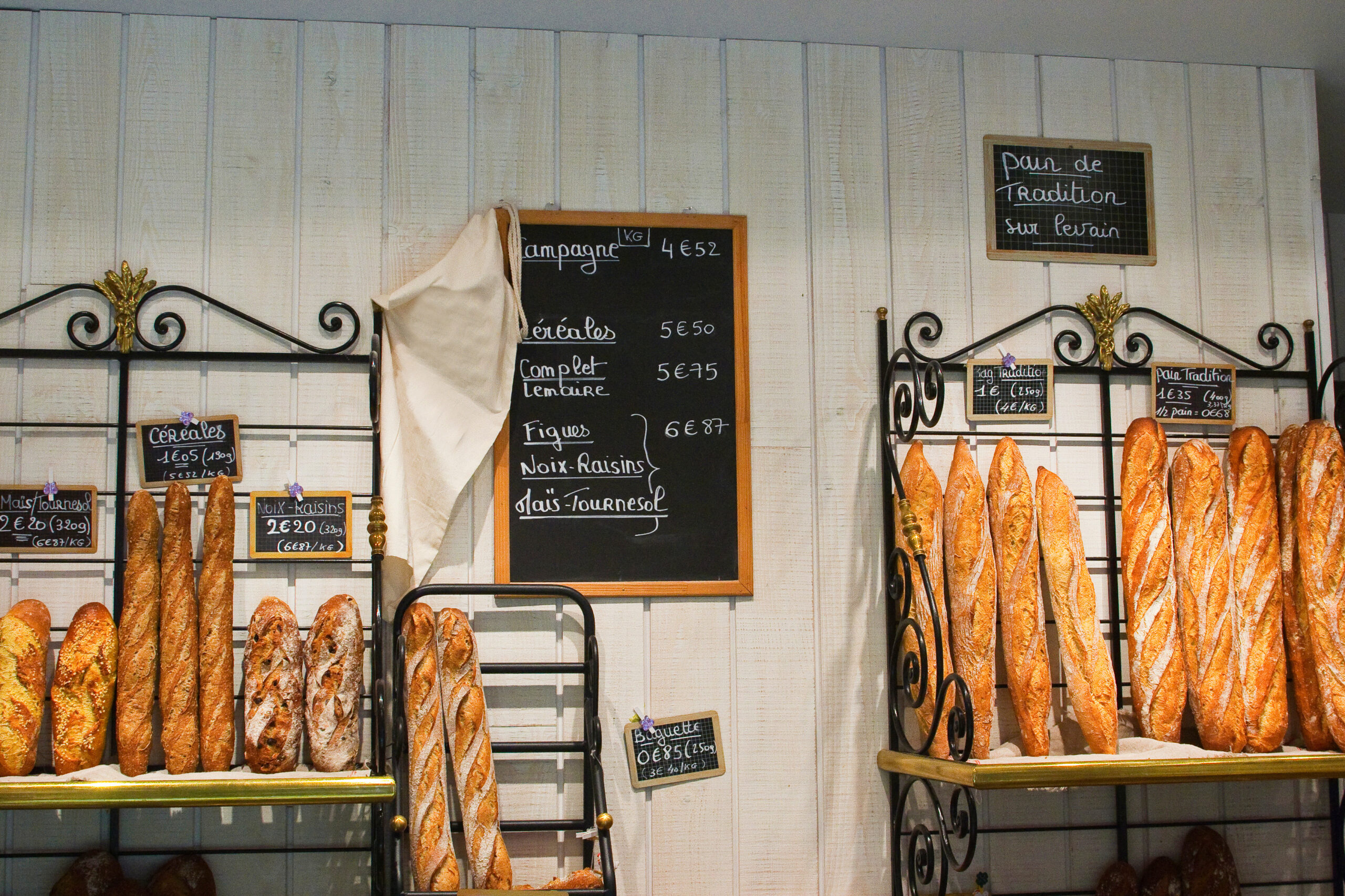Rows of baguettes hanging on black iron racks in a bakery, with chalkboard menus on the wall behind them.