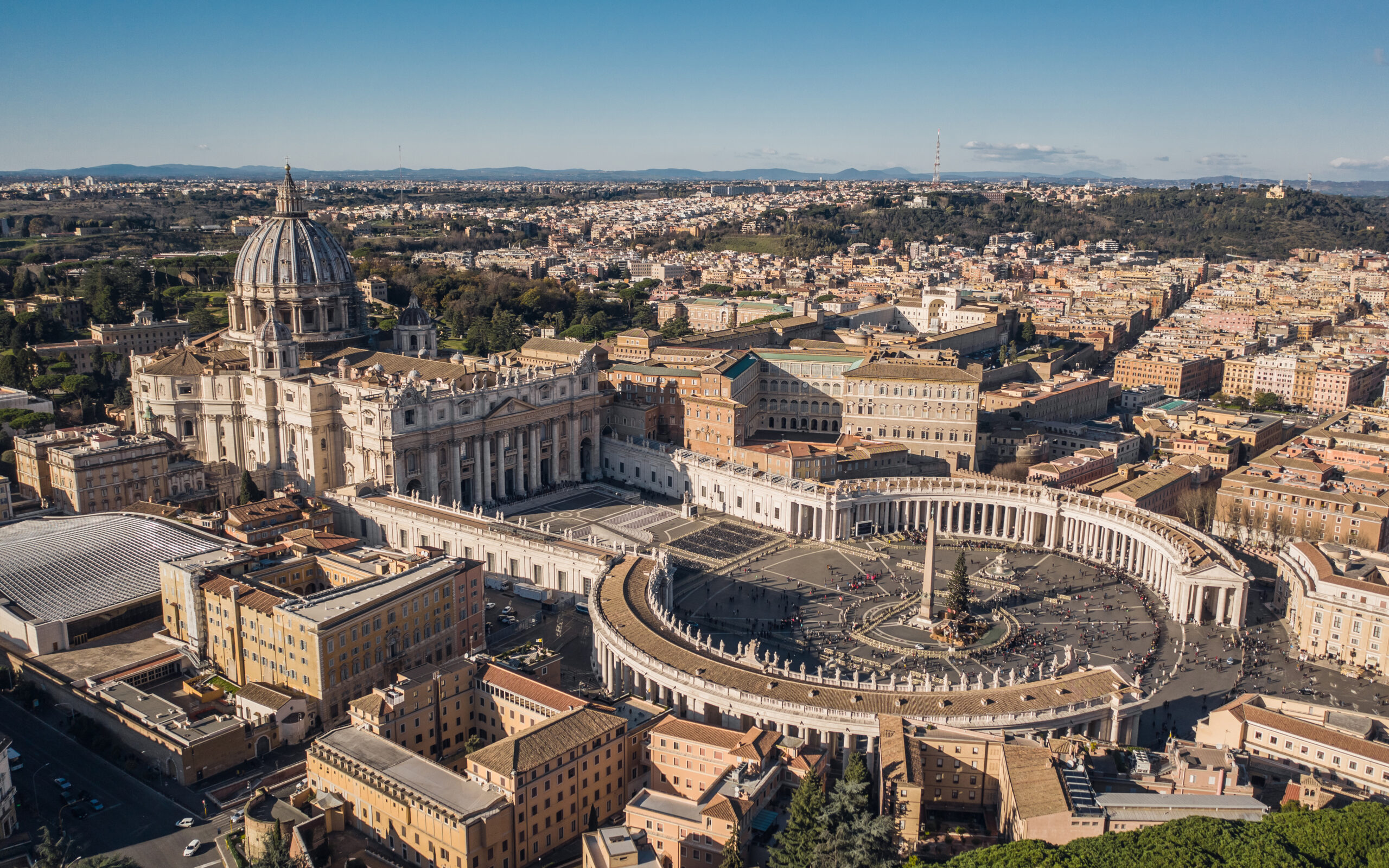 Aerial view of St. Peter's Basilica with its large dome and the surrounding colonnade, overlooking Vatican City and Rome under a clear blue sky.