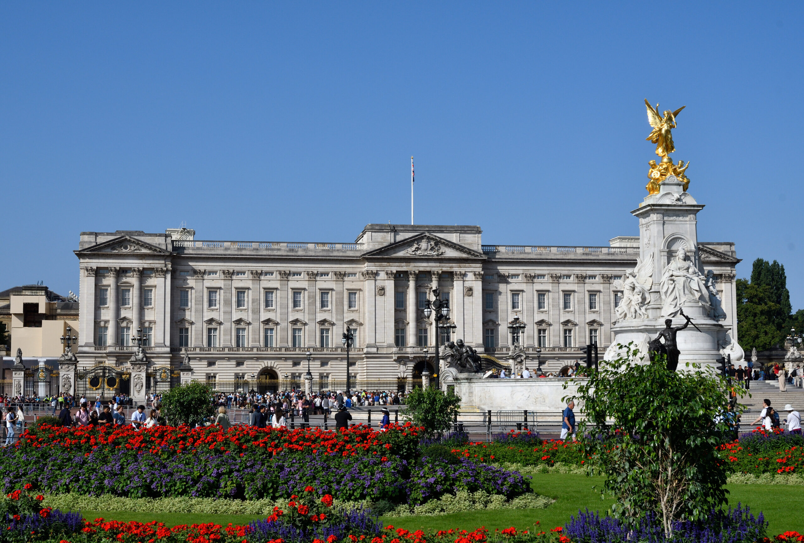 Buckingham Palace with the gilded Victoria Memorial statue to the right and crowds in front, with colorful flower beds in the foreground down the lawn.