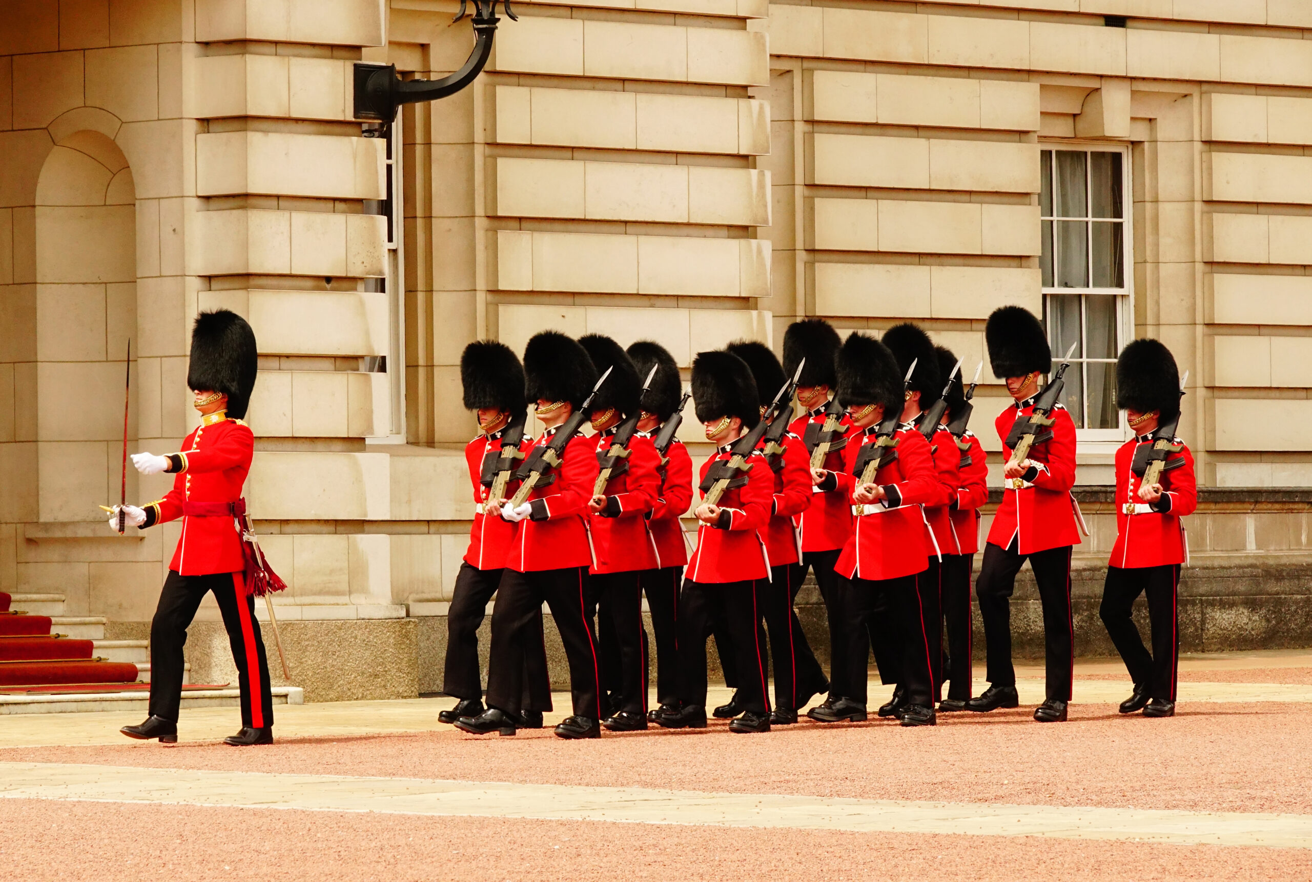 Ceremonial guards in red uniforms with tall bearskin hats marching in formation, rifles at the ready, outside a stone building.