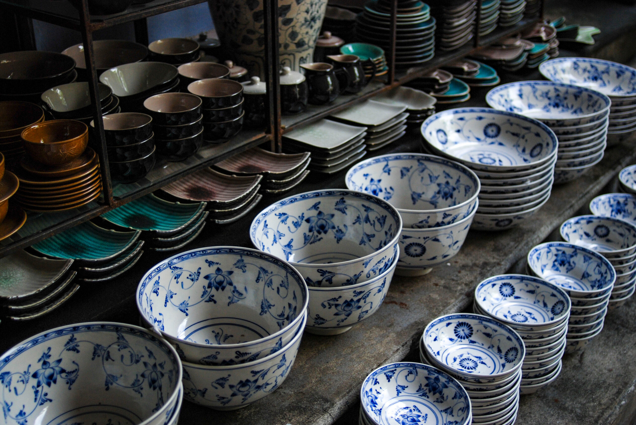 Stacks of blue-and-white porcelain bowls and plates on display shelves in a pottery market stall, with various ceramic pieces nearby.