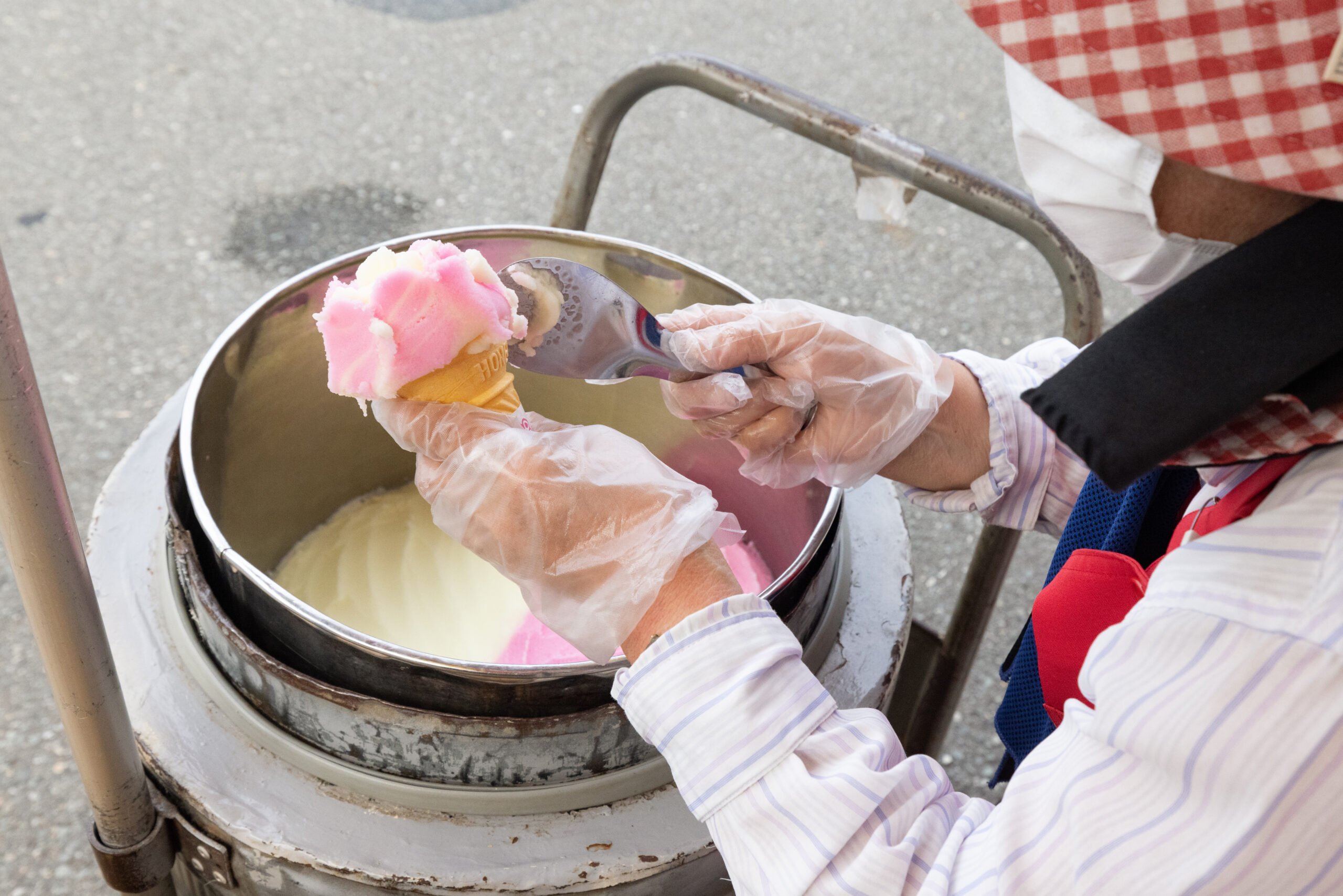 Gloved hands scoop pink ice cream from a metal tub at an outdoor cart.