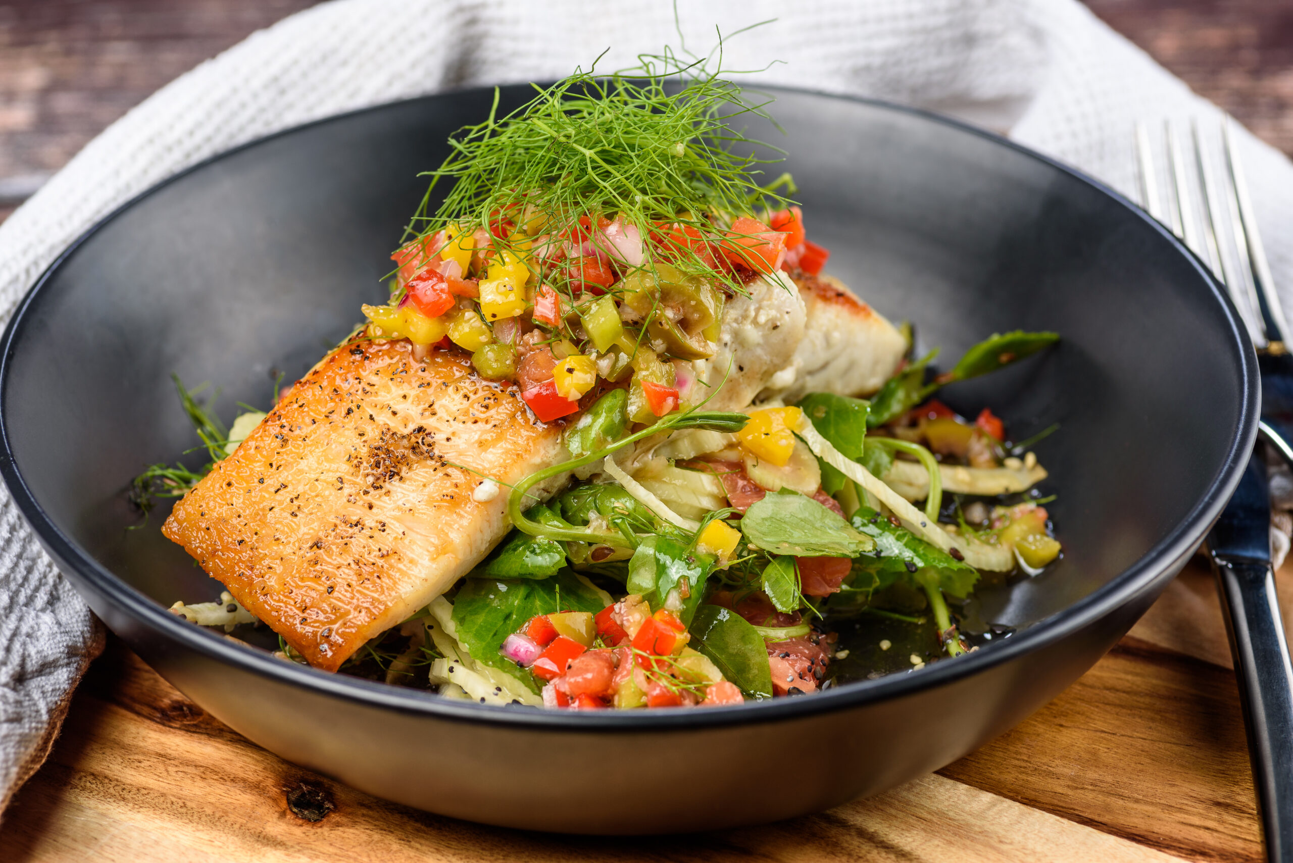 Pan-seared salmon fillet topped with colorful salsa and dill on a bed of greens in a black bowl.