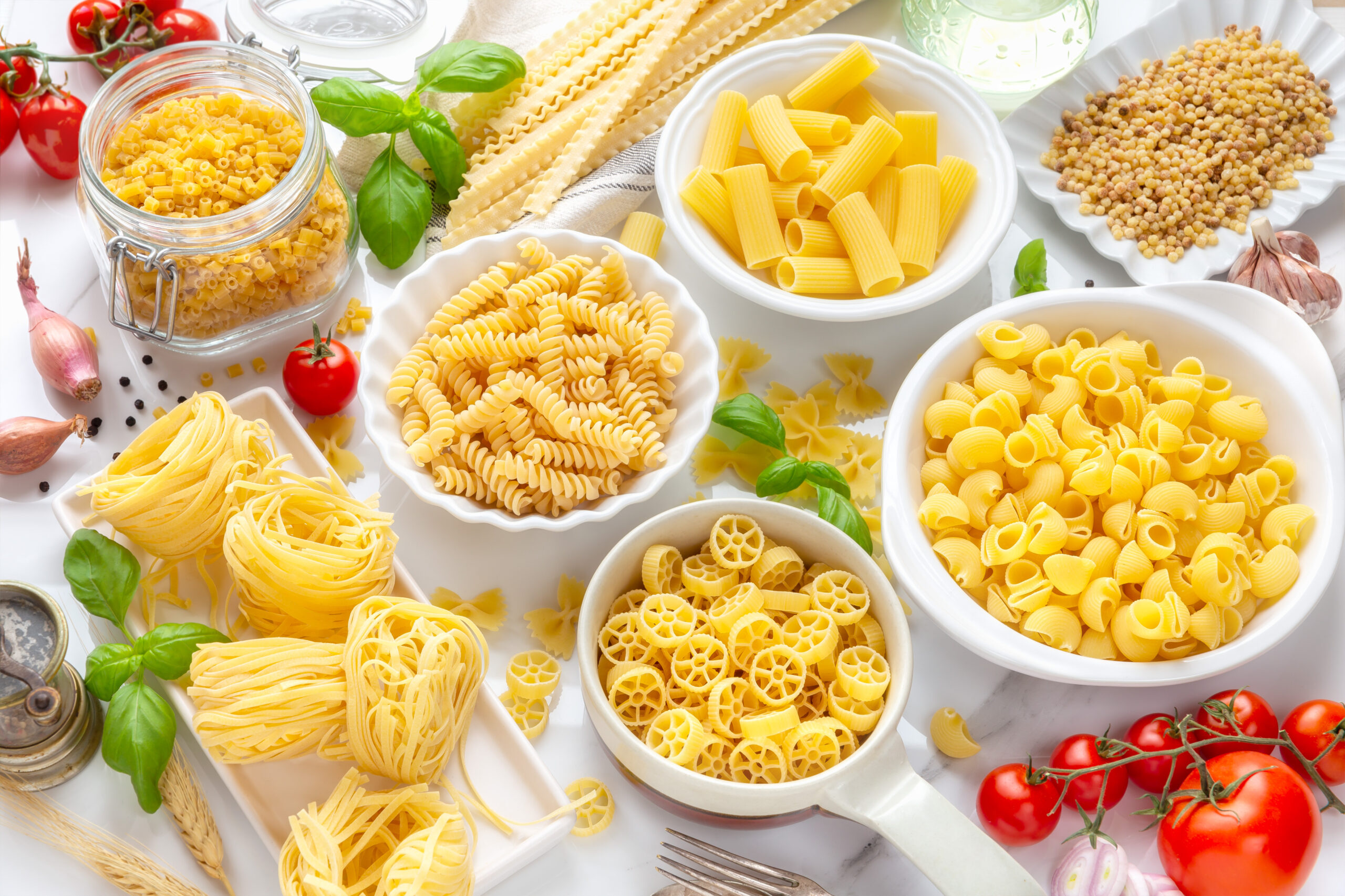 Assortment of uncooked pasta shapes in bowls and jars with tomatoes and basil on a white table.