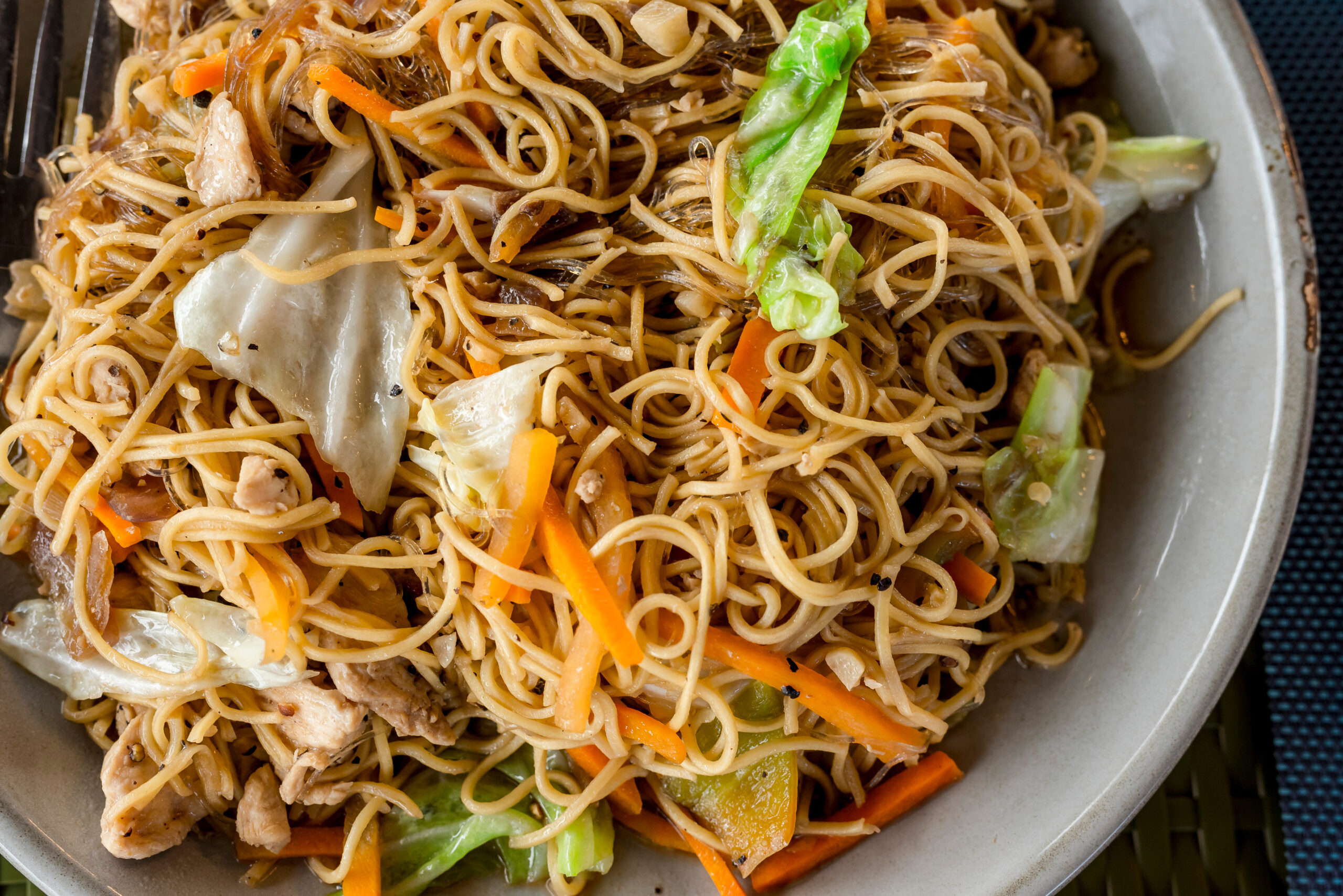 Stir-fried noodle dish with chicken pieces, carrots, cabbage, and green vegetables in a skillet