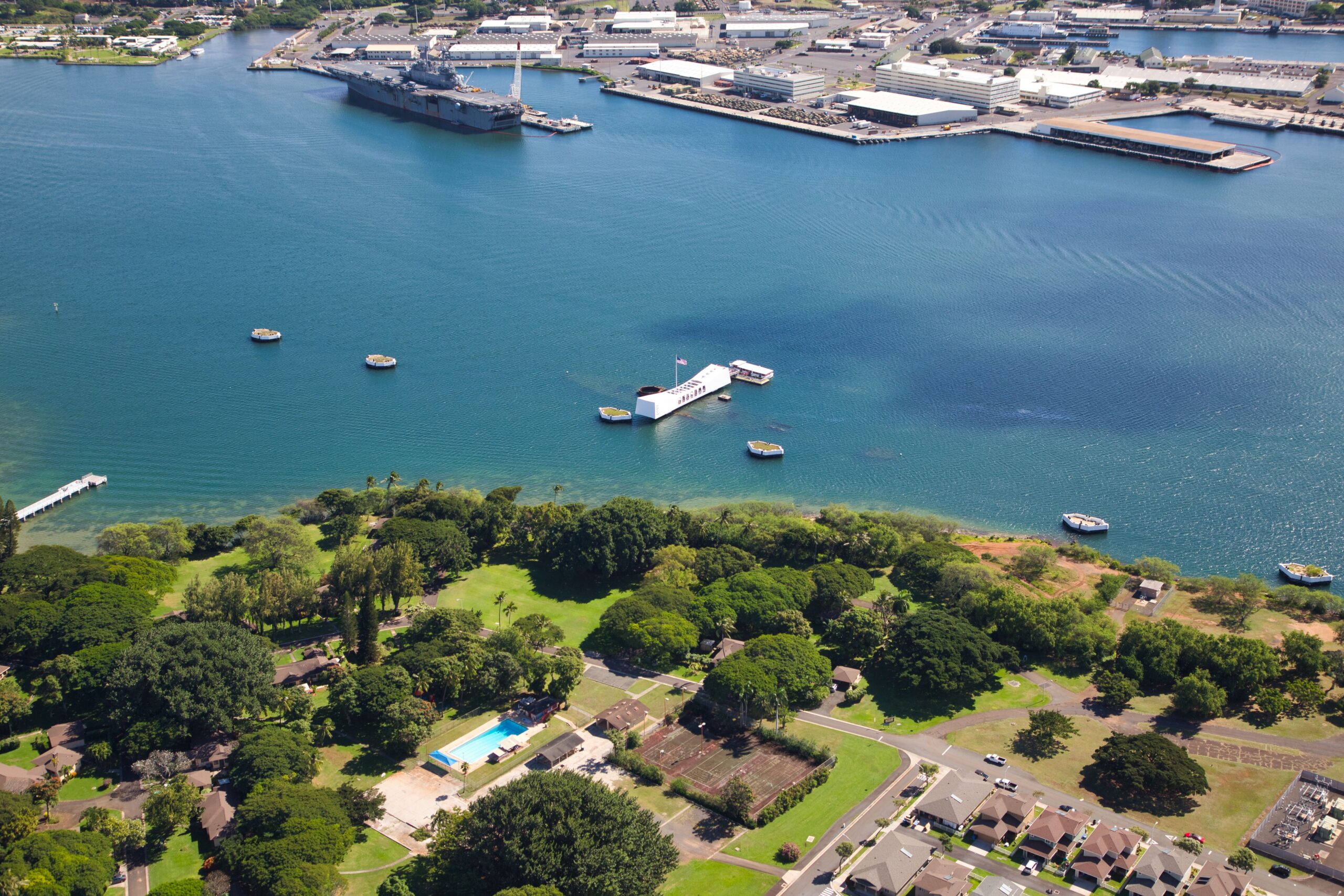 Aerial view of a harbor with a large ship docked at a pier and blue water, surrounded by industrial buildings and docks.