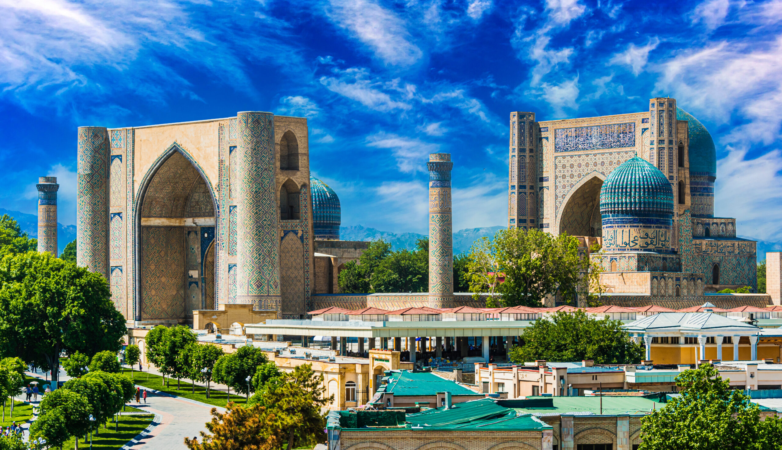 Historic Islamic complex with blue-tiled domes, tall arched gateways, and intricate mosaics under a bright blue sky.