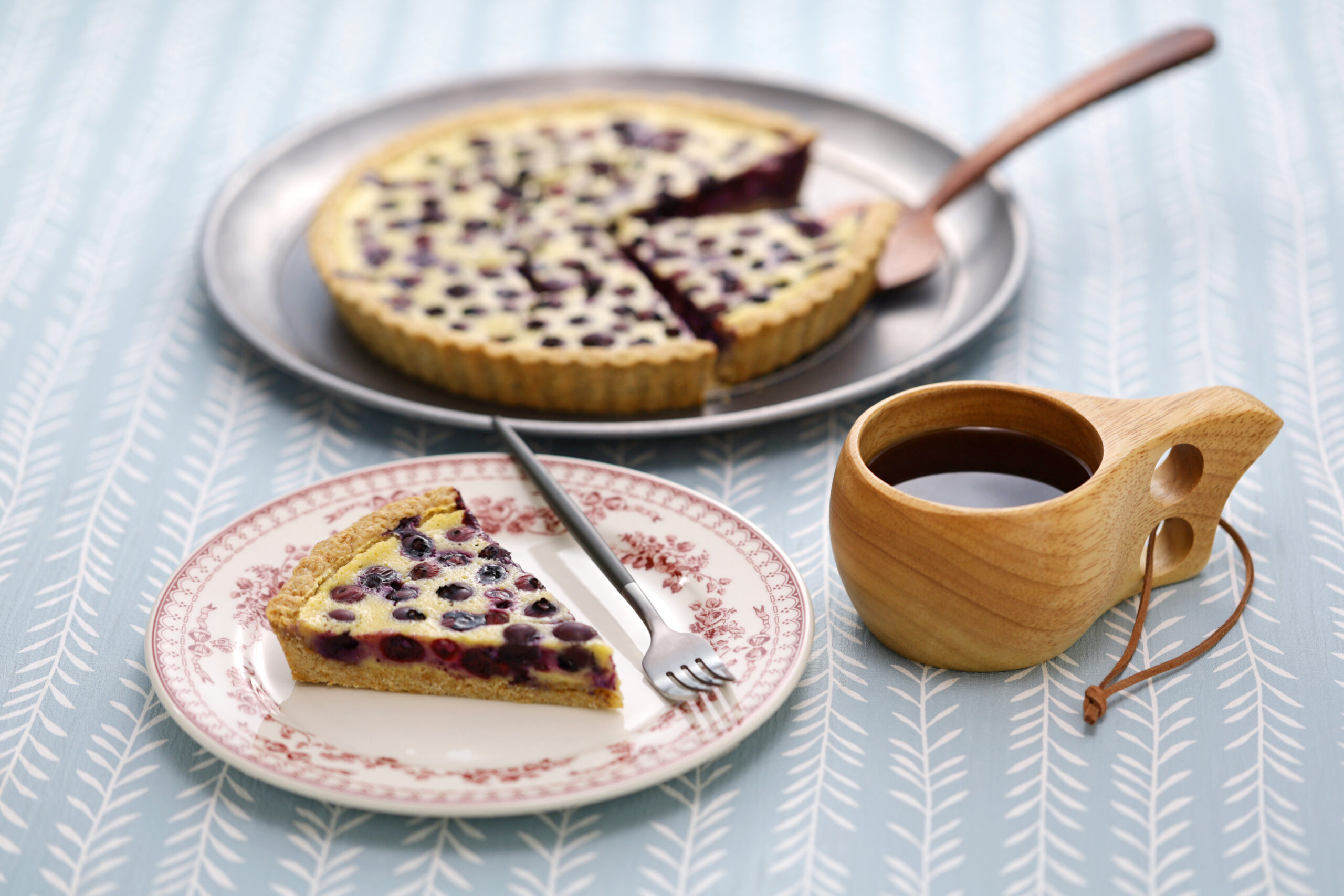 Slice of blueberry tart on a patterned plate with a fork, set on a pale blue tablecloth with a chevron pattern.