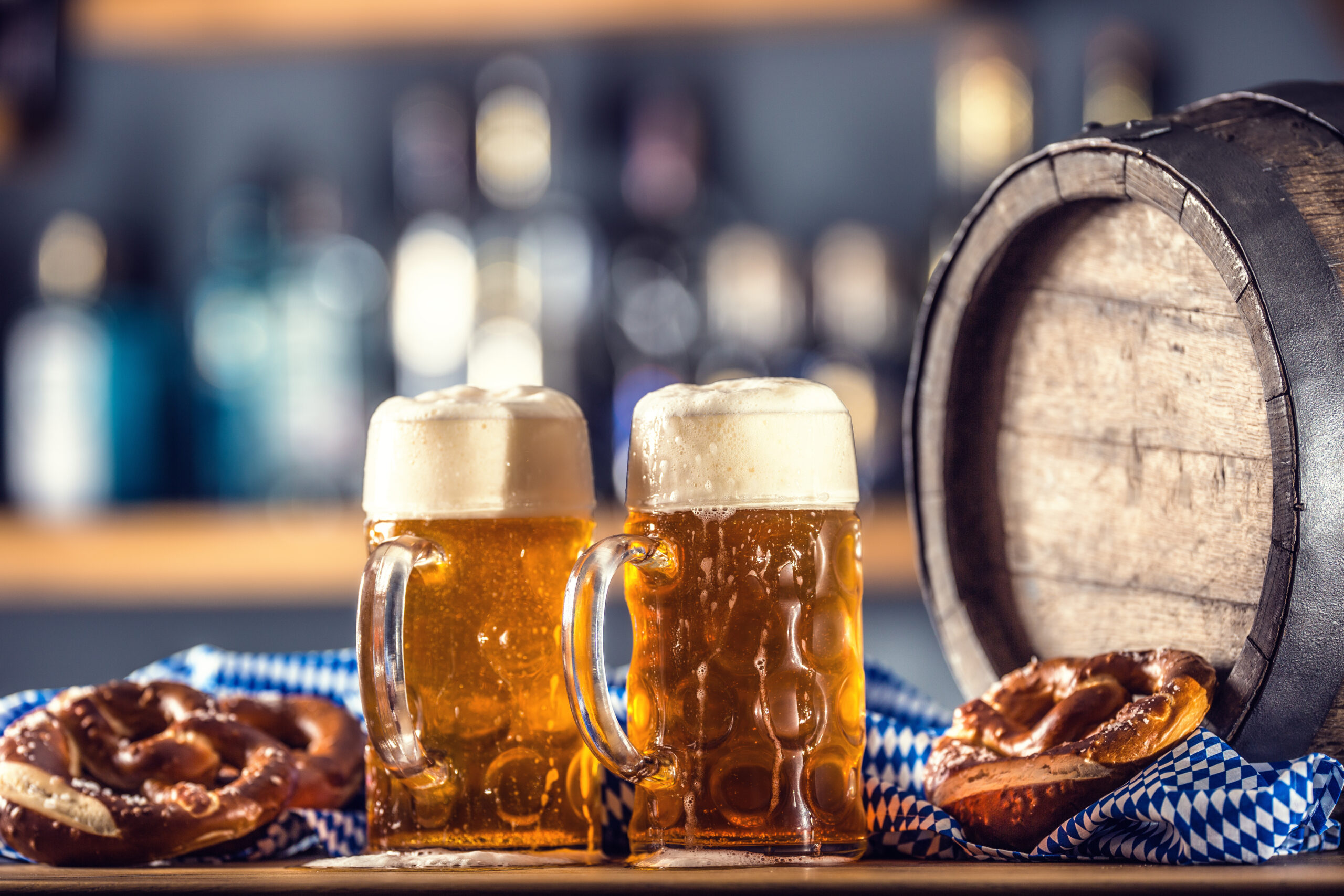 Two frothy beer mugs sit on a wooden table, with a large wooden barrel in the background and pretzels on a blue-and-white checkered cloth nearby.