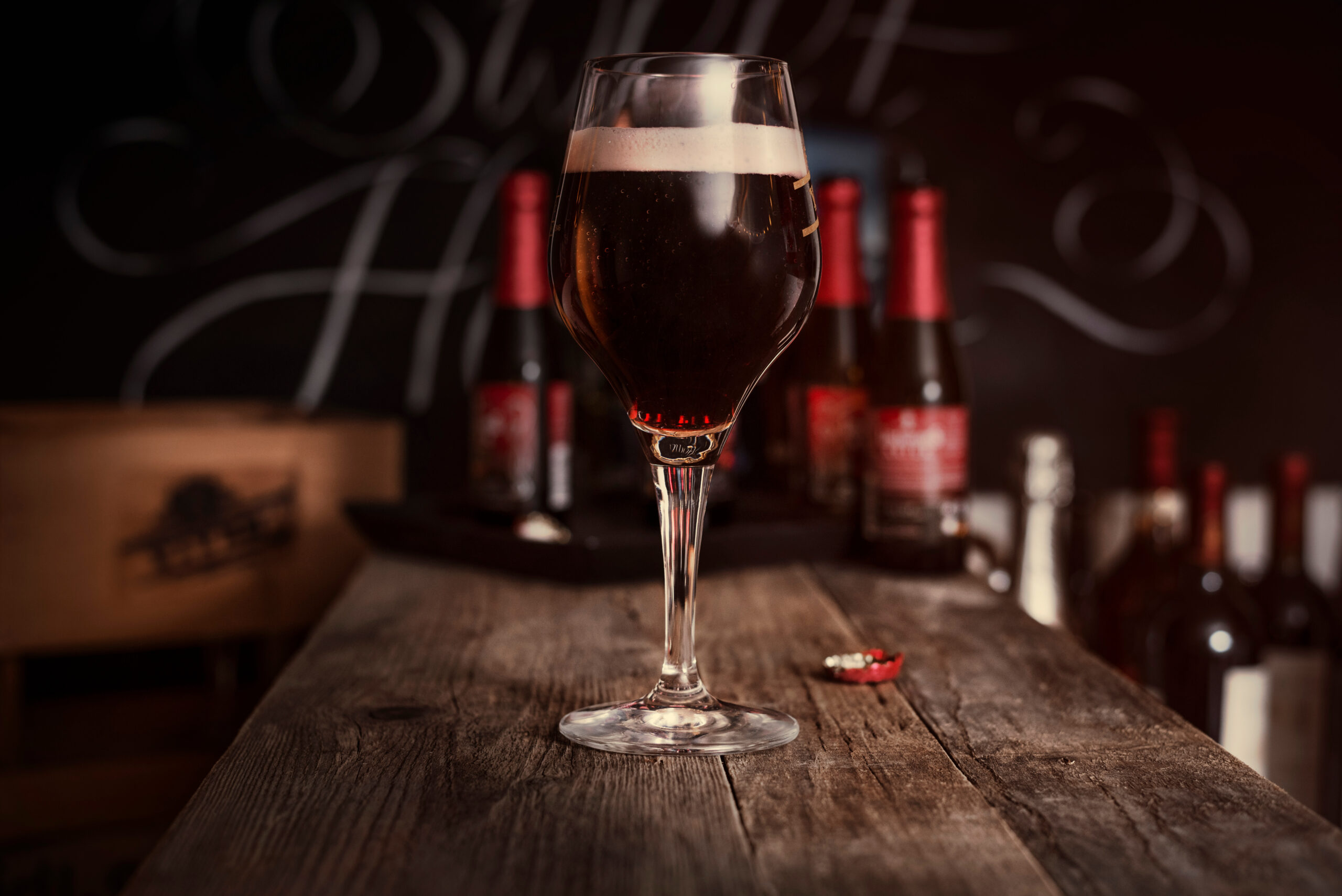 Dark beer in a tall stem glass with a creamy head on a rustic wooden bar, blurred bottles in the background.