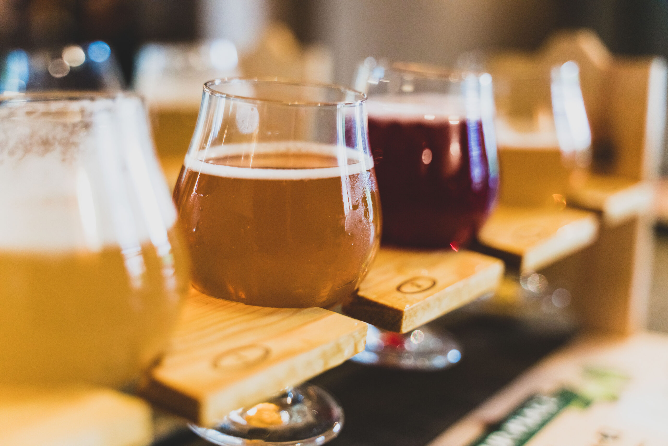 Close-up of several beer glasses on a wooden bar, with an amber ale in the foreground and darker beers blurred in the background.
