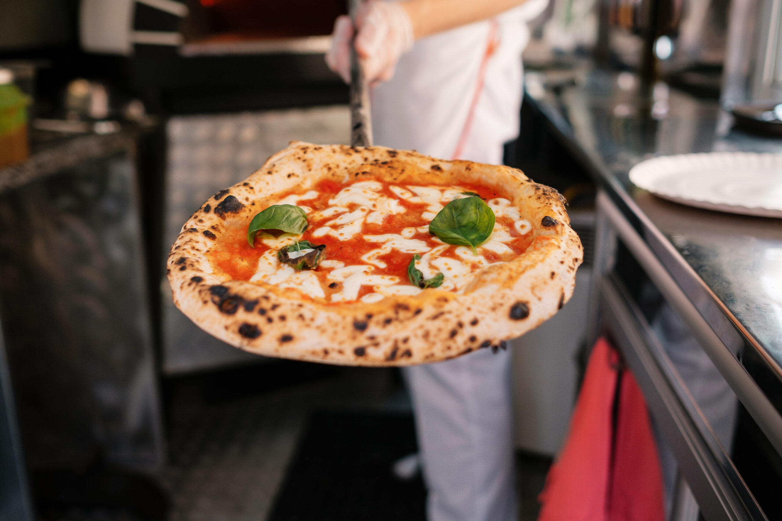 Chef in white holds a hot, fresh pizza toward the camera in a busy kitchen, basil leaves visible on top.