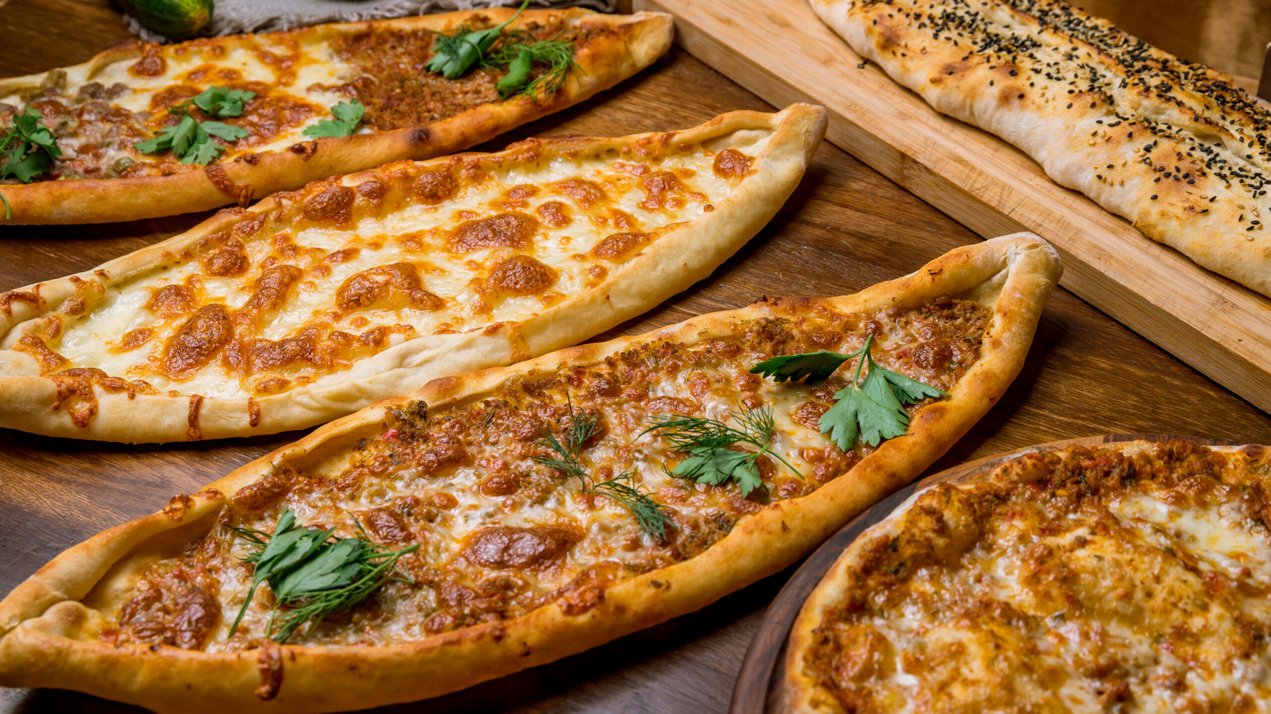 Assorted pizzas on a wooden table, topped with cheese and fresh herbs, plus a sesame-seed bread roll on the side.