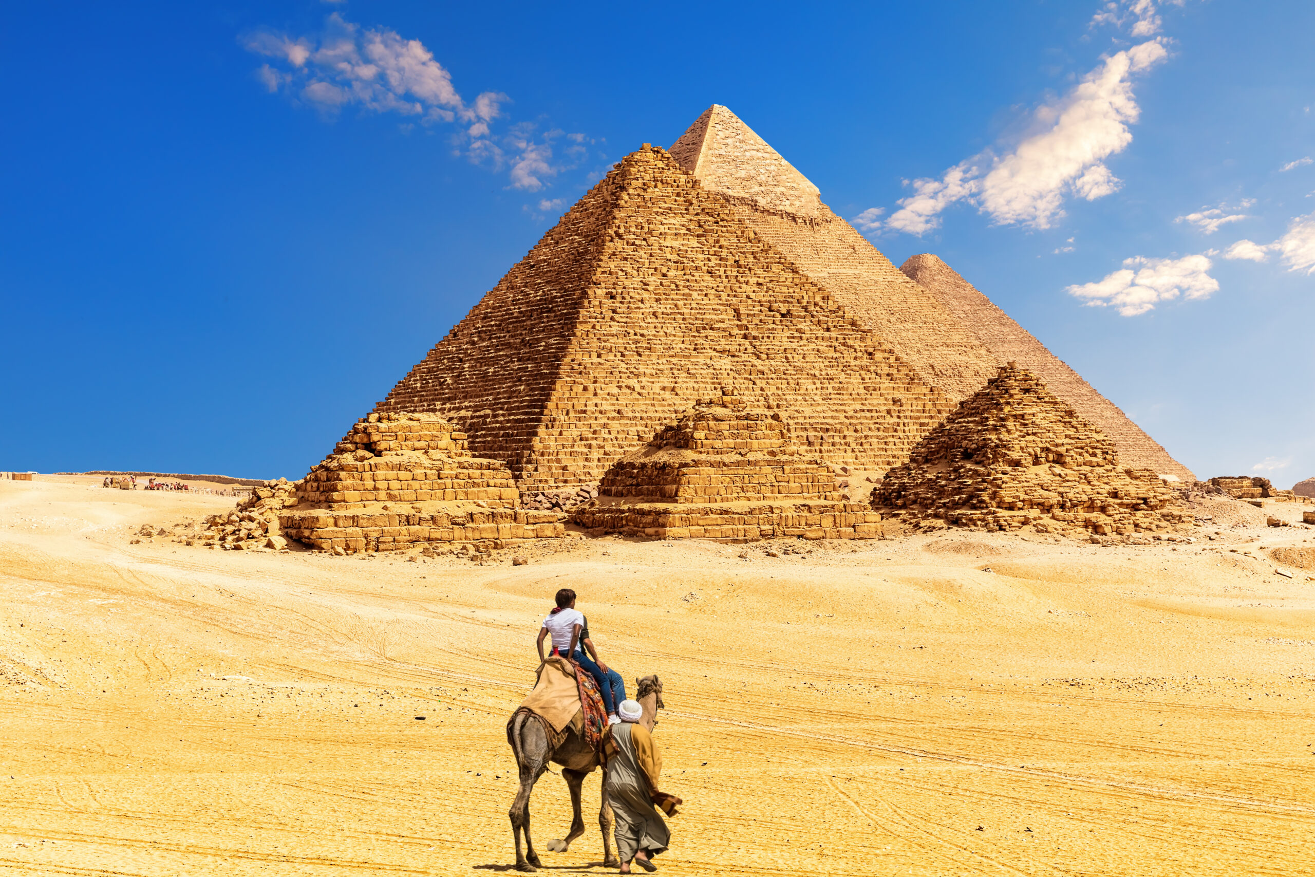 Tourists ride camels in the foreground with the pyramids of Giza under a bright blue sky in the background due to desert scenery.