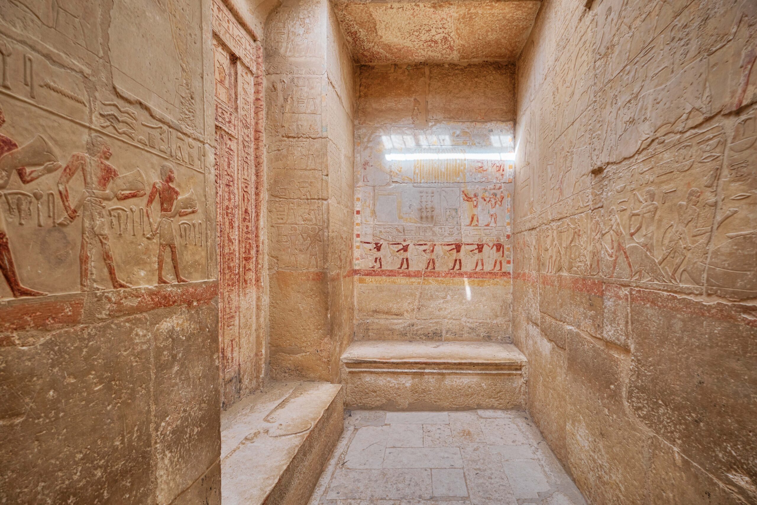 Stone corridor of an ancient Egyptian tomb with hieroglyphic carvings and painted reliefs along the walls, fading light at the far end.