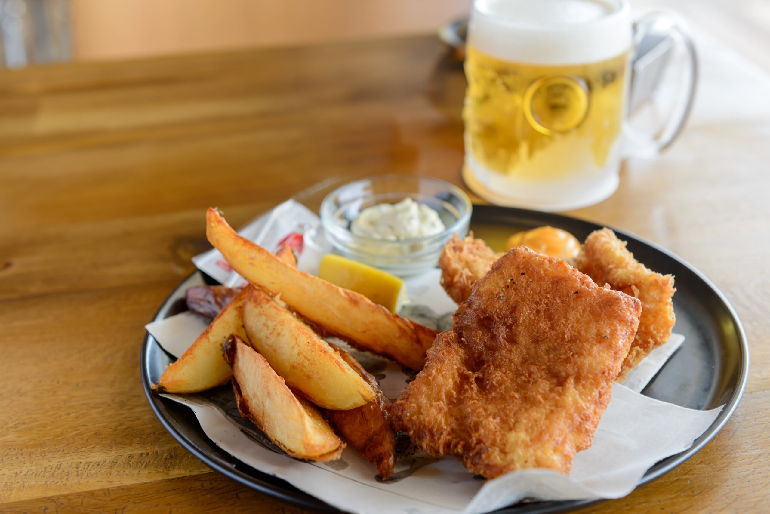 Plate of crispy fried fish and thick-cut fries with dipping sauce and a lemon wedge on parchment, on a wooden table with a beer mug in the background