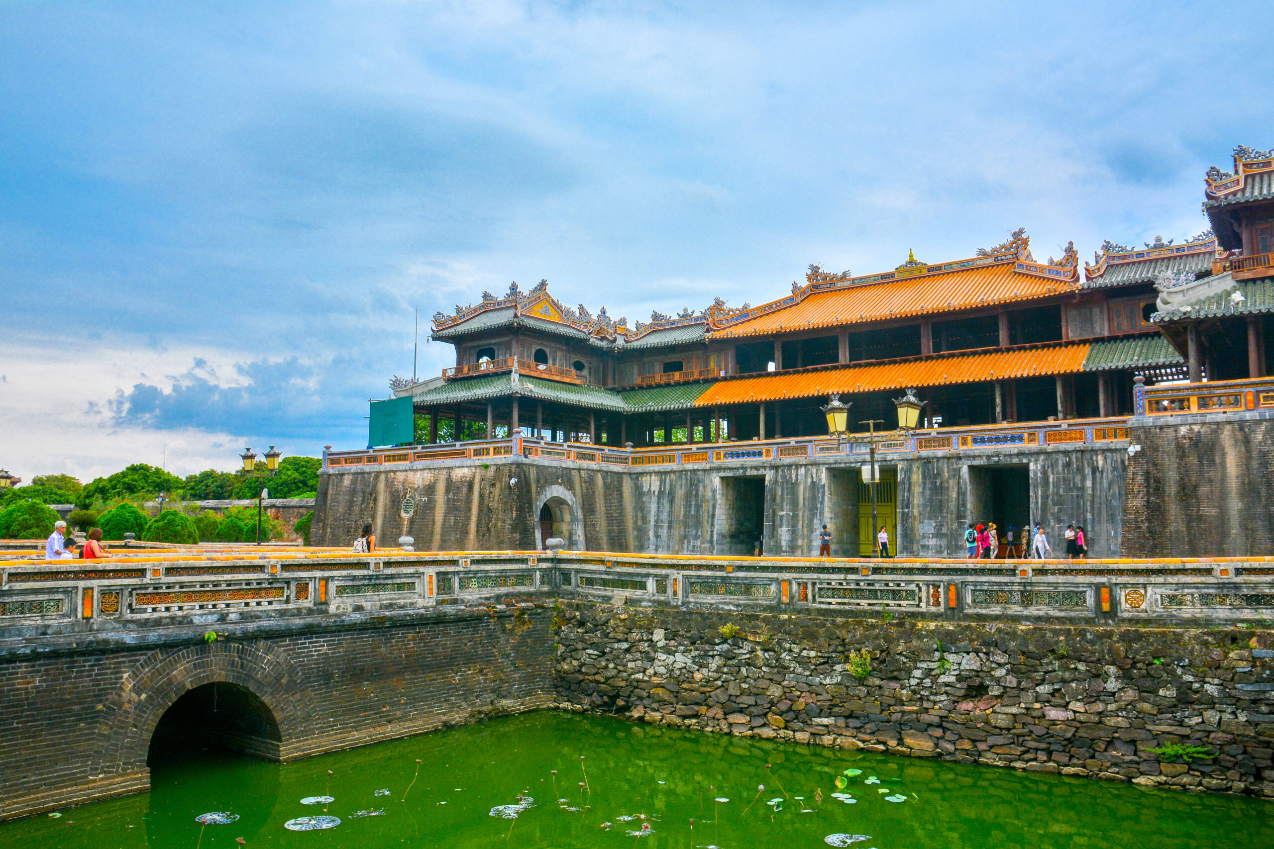 Stone fortress with colorful tiled roofs and decorative eaves along a moat; visitors stroll the walkway.