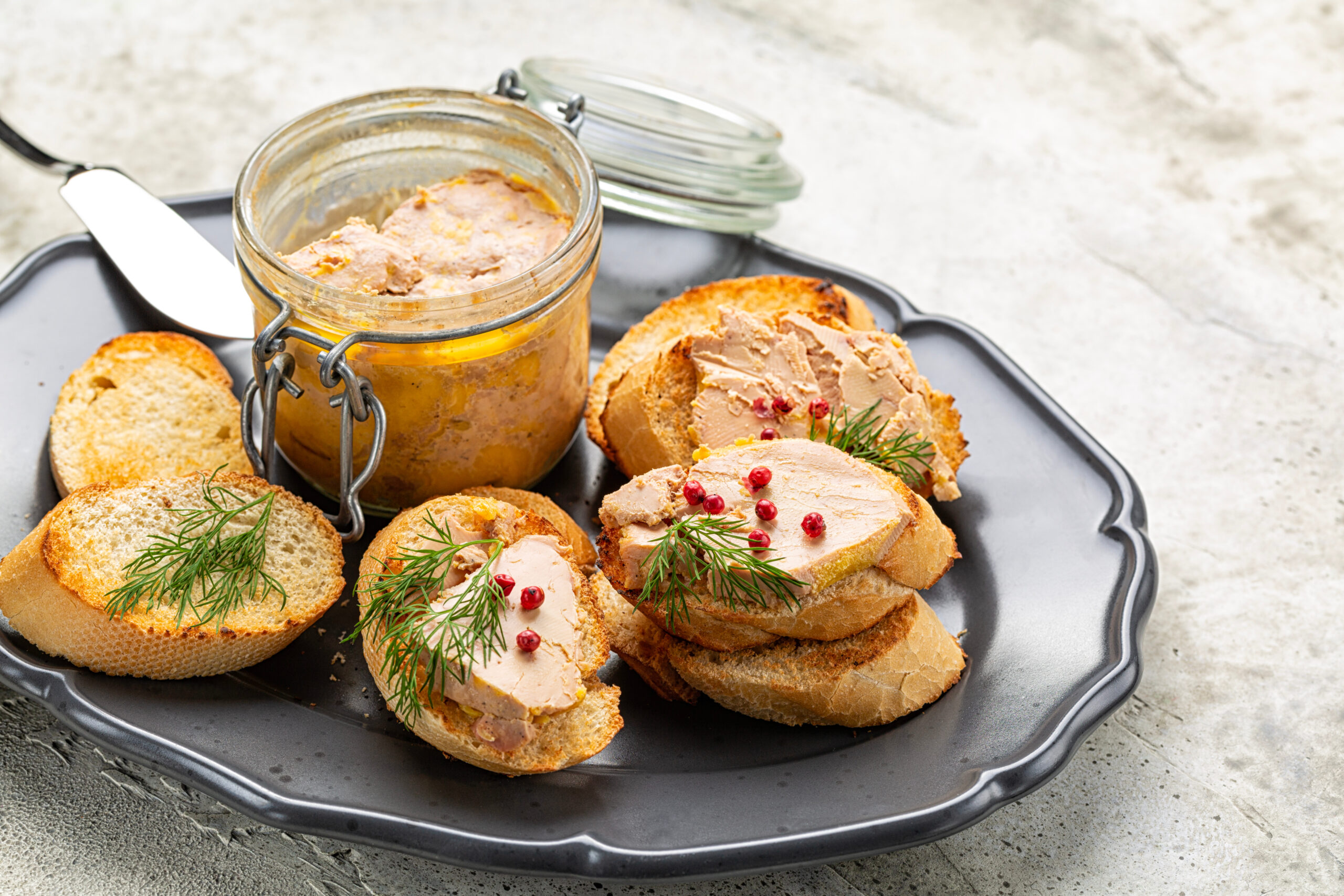 Jar of pâté in a glass container with bread rounds topped with pâté, dill sprigs, and pink peppercorns on a serving tray
