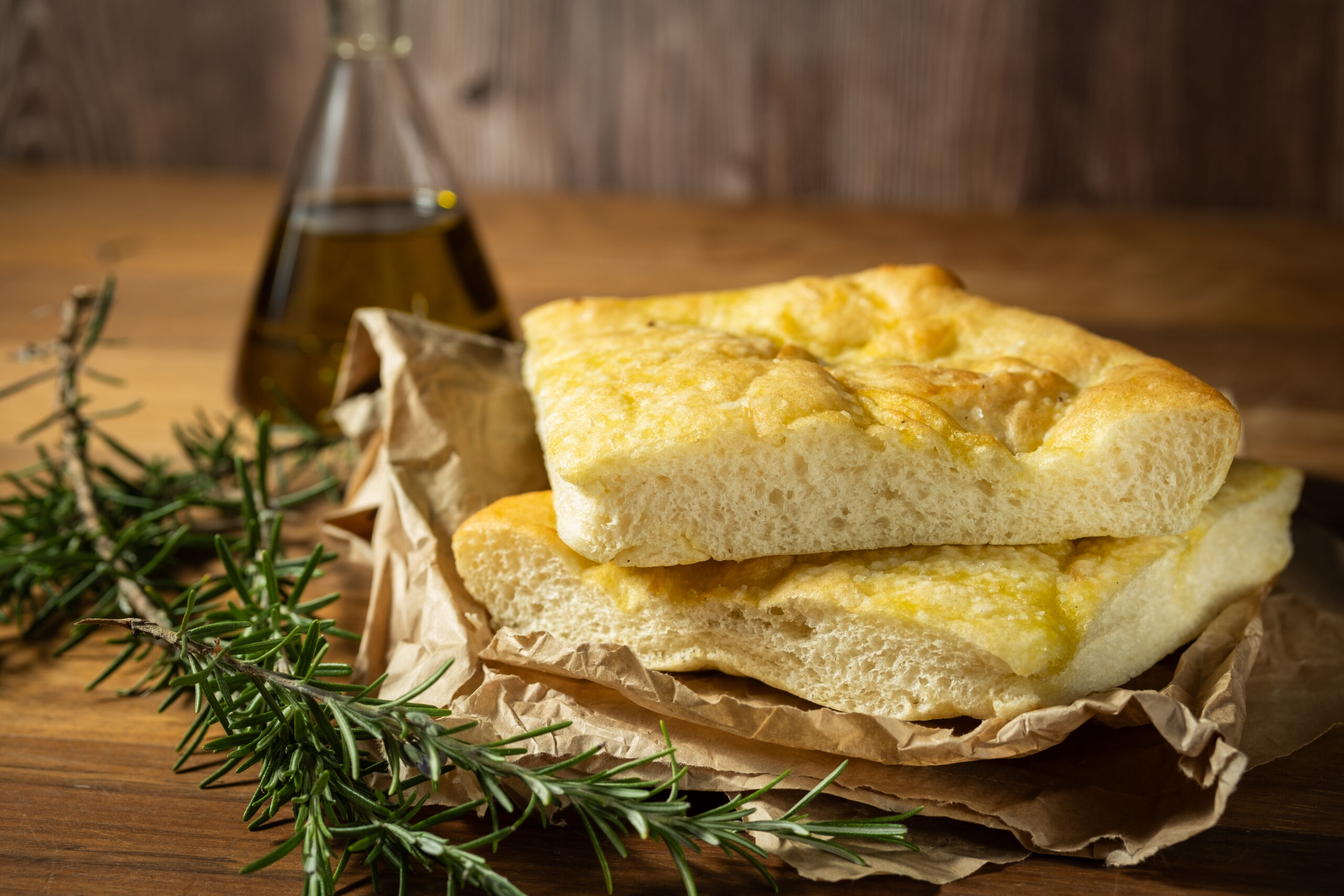 Slices of focaccia bread with a golden crust on parchment, rosemary sprigs in foreground and an oil bottle in the background on a wooden table.