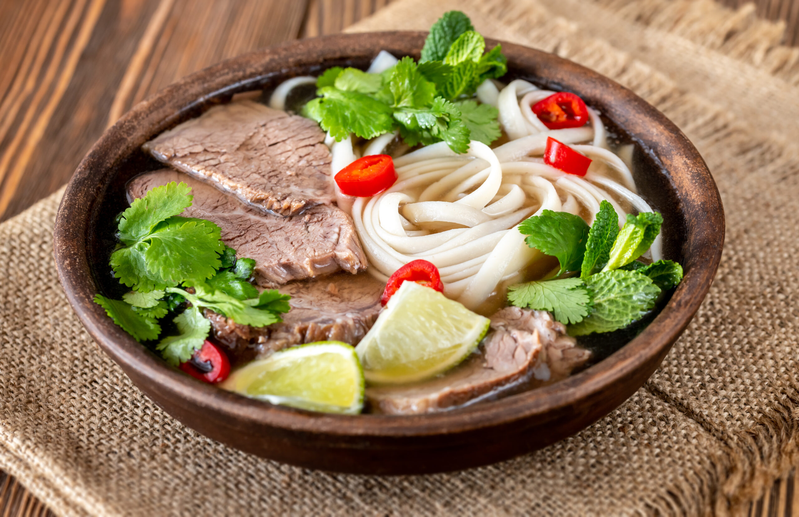Beef noodle soup in a rustic wooden bowl topped with fresh cilantro, lime wedges, and sliced red chilies on a burlap surface