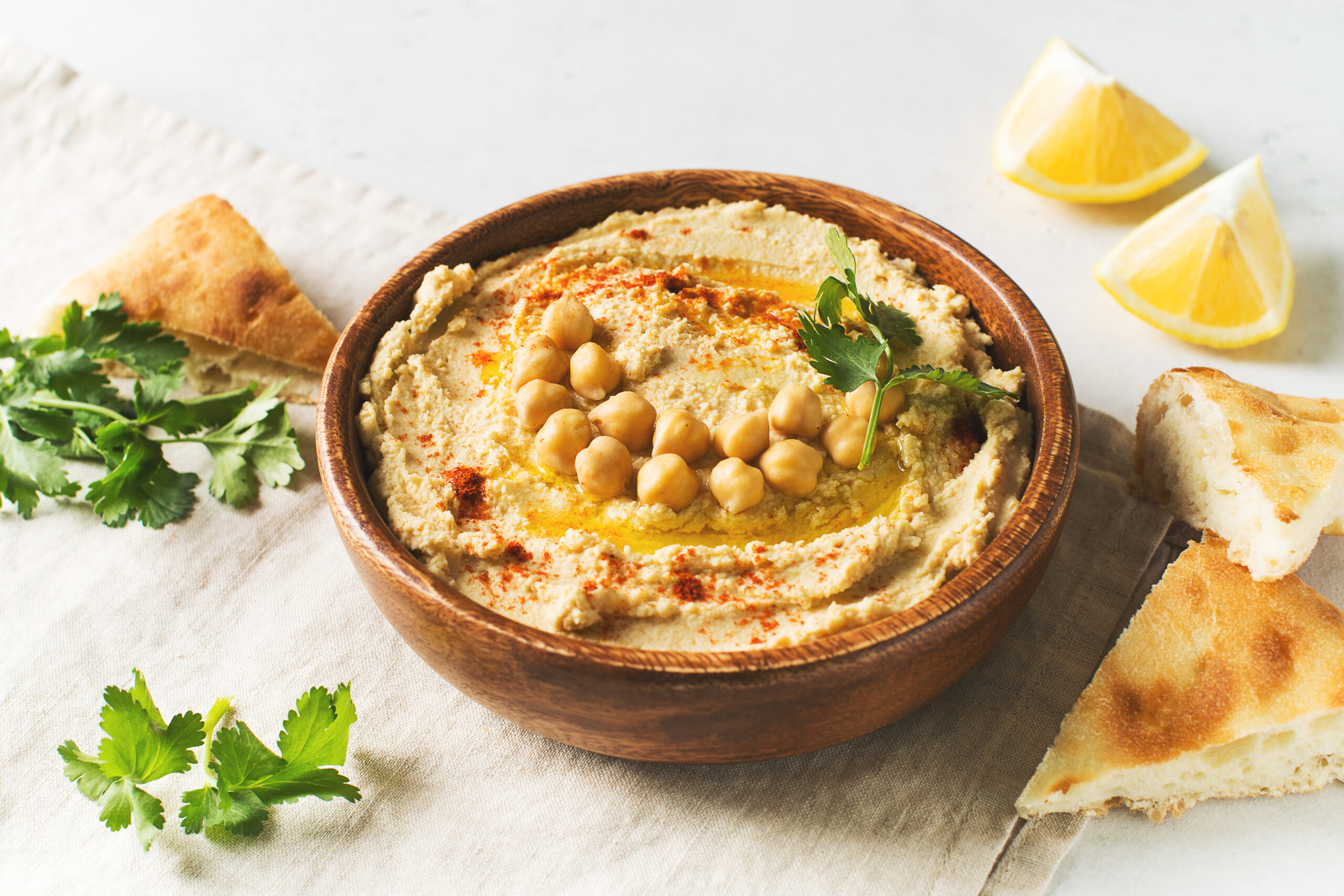 Hummus in a wooden bowl topped with chickpeas, olive oil, paprika, and parsley, with pita bread and lemon wedges nearby.