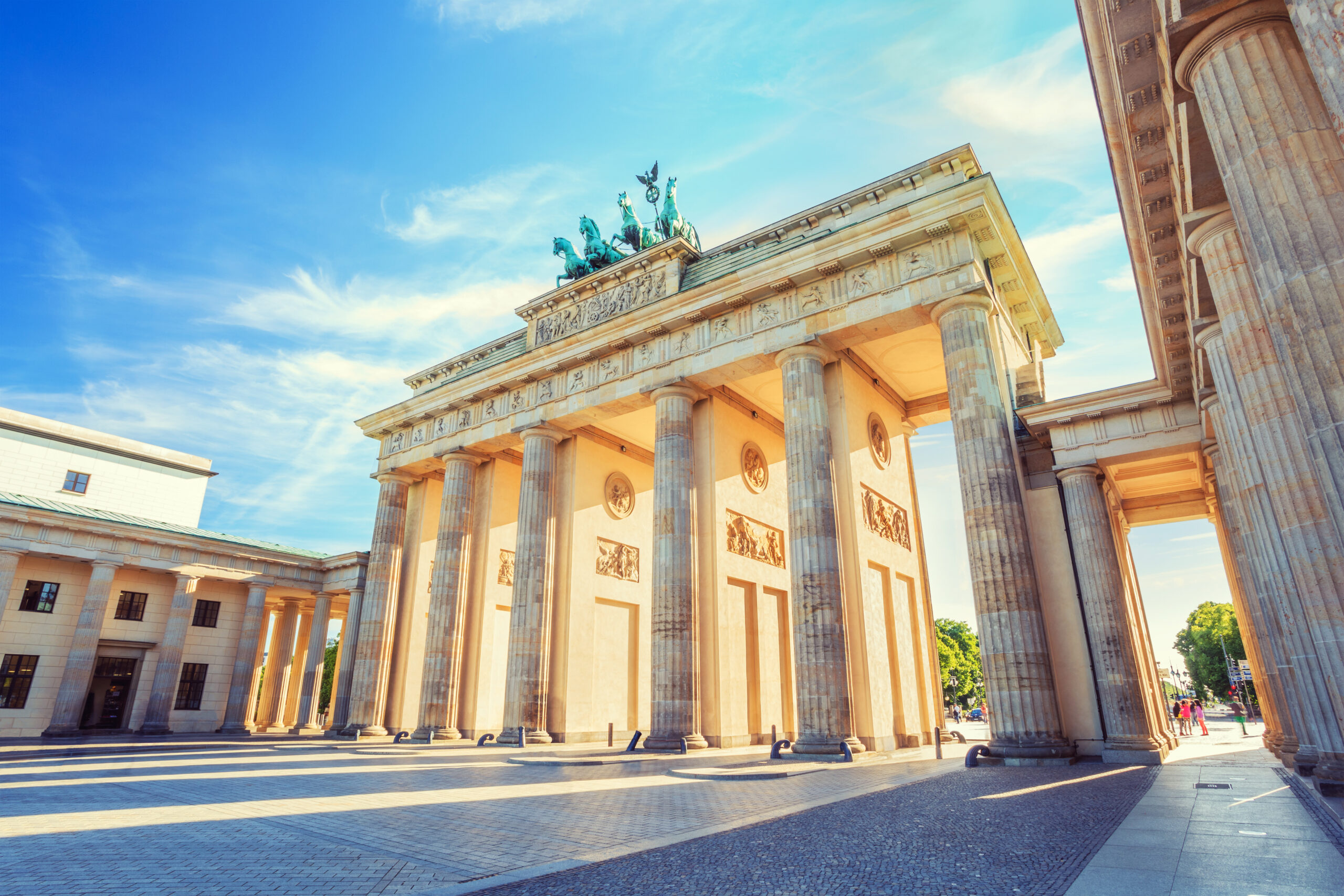 Brandenburg Gate–style neoclassical arch with 12 columns and a chariot statue on top against a bright blue sky.